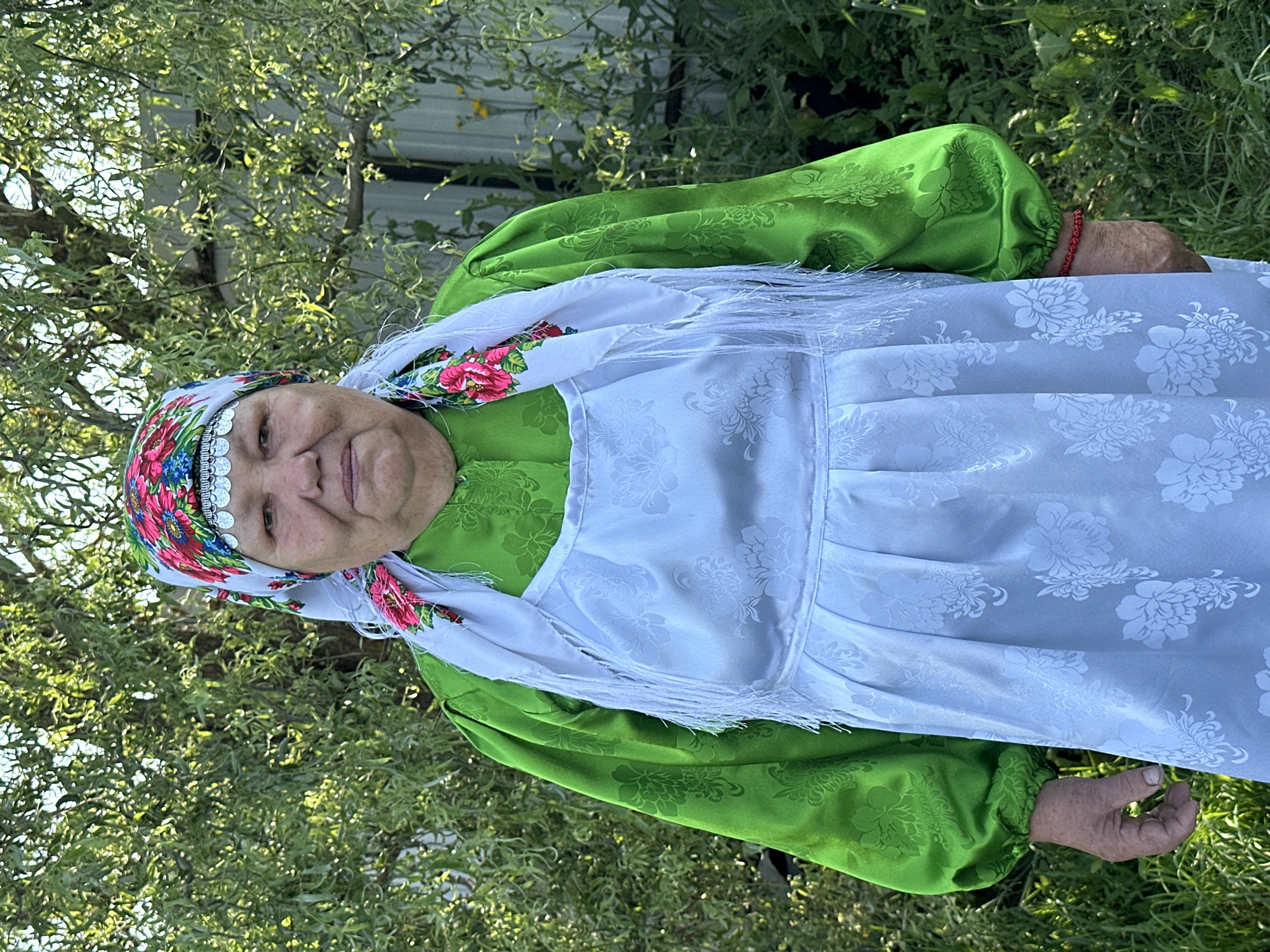 An elderly woman stands outdoors, facing forward, in what appears to be a village setting. She wears a vibrant green long-sleeved blouse with textured floral patterns, a white apron with a similar floral design, and a colorful headscarf adorned with flowers and silver embellishments. A white scarf with fringed edges is draped around her neck and shoulders. She has a red bracelet on her right wrist. The background consists of lush green foliage and trees, suggesting a natural or garden environment. A portion of a metal fence and some yellow flowers are visible behind her. The lighting indicates it is daytime. The location is specified as Sredneye Kechevo, Russia.