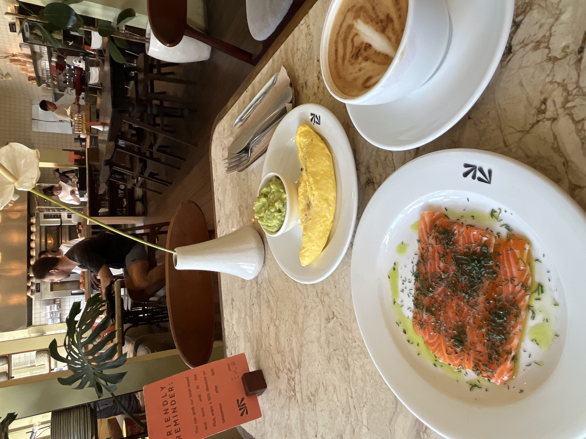 A table in a bright, modern cafe setting in North Kuta, Indonesia, displays a breakfast spread. In the foreground, a white plate holds a serving of thinly sliced salmon sprinkled with dill and drizzled with olive oil. Adjacent to it, another white plate contains a folded omelet and a small bowl of mashed avocado. A white cup and saucer with a latte art-decorated coffee sit to the right. A white vase with a delicate flower stands behind the food. In the background, a man is seated at a table, looking towards a laptop. Other patrons and staff are visible in the cafe, with shelves of products and kitchen equipment lining the walls. A vibrant green plant with large leaves is positioned to the left of the scene.