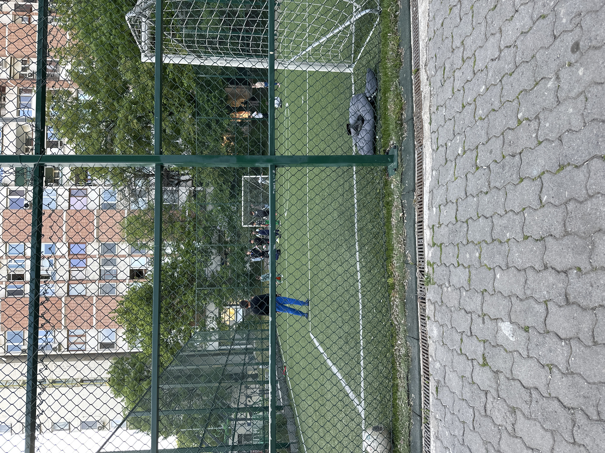 The image, taken in Sofia, Bulgaria, depicts a small, fenced-in artificial turf soccer field. A chain-link fence encloses the field, with a visible building featuring a grid of windows beyond the fence. The field itself has white lines marking the boundaries and contains two soccer goals at either end. Several people are visible on the field, some standing near one of the goals. One person wearing blue pants is standing closer to the center of the field. A gray bag rests along the edge of the field near the fence. The ground adjacent to the fence is paved with interlocking gray stones. A metal drainage grate runs along the edge of the paving. Trees are visible in the background. The image is taken through the fencing, partially obscuring the scene.