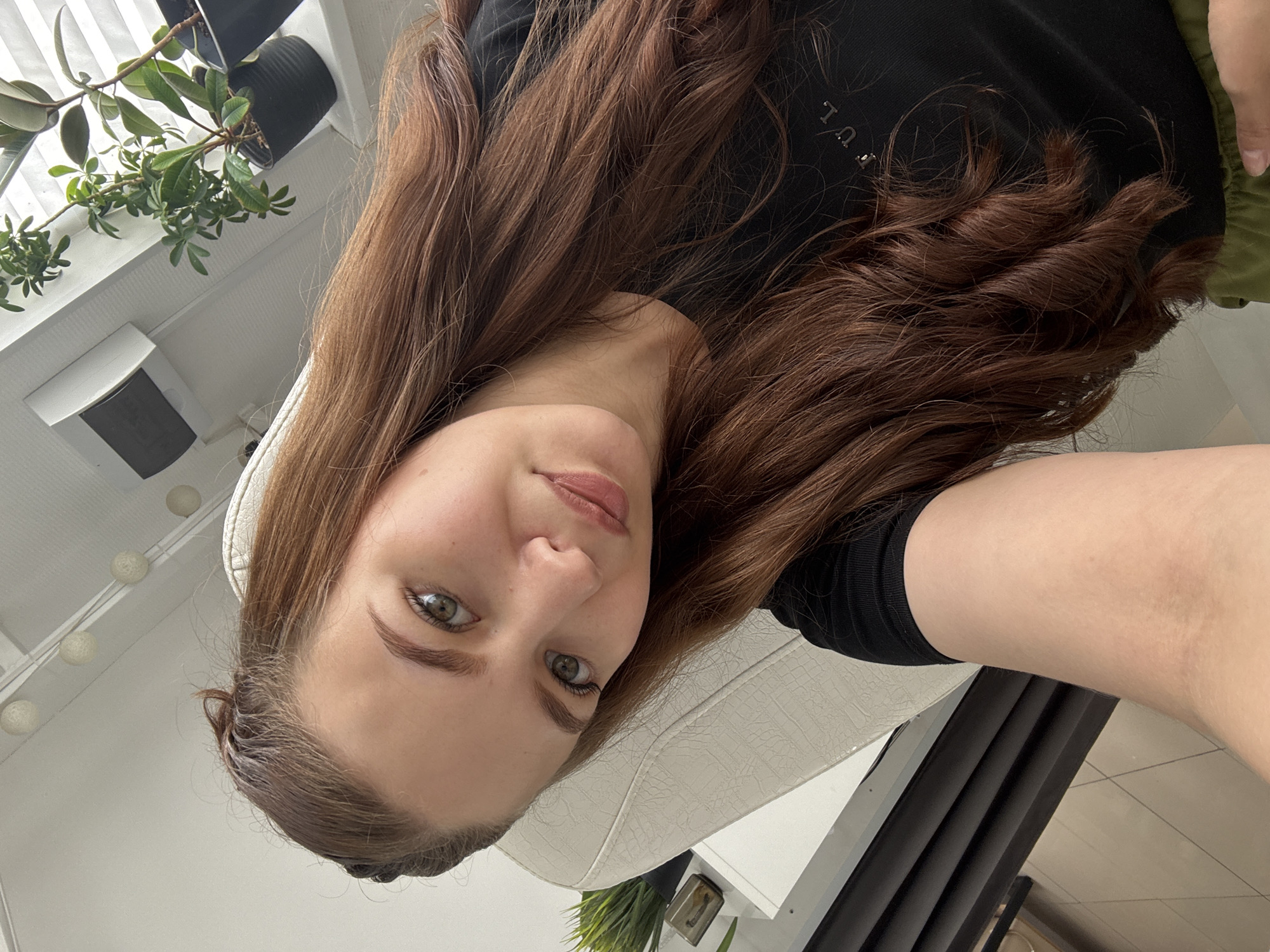 The image shows a selfie of a young woman with long, auburn hair, taken indoors. She is wearing a black t-shirt. Her hair is partially braided at the top. She is positioned in front of a white chair, and a desk with plants and blinds is visible in the background. The lighting suggests an indoor setting, possibly near a window. The image is taken from a slightly high angle, looking down at the subject. The location is not specified in the image itself, and there is no information linking it to Chaikovsky, Russia.