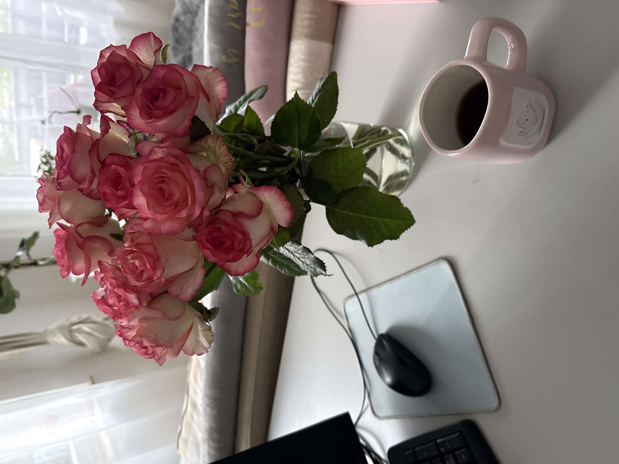A bouquet of pink and white roses sits in a clear glass vase on a white desk. The roses have dark green leaves and some stems. A black computer mouse rests on a light blue mousepad to the left of the roses. In front of the mousepad is a black computer keyboard. To the right of the roses is a pink mug filled with a dark liquid, possibly coffee. The mug has a white square decal with a cartoonish design on it. In the background, stacks of books or rolled fabric are visible. Sheer white curtains hang in the background, allowing natural light to illuminate the scene. The overall impression is a quiet and serene desk setting in Bad Sachsa, Germany.