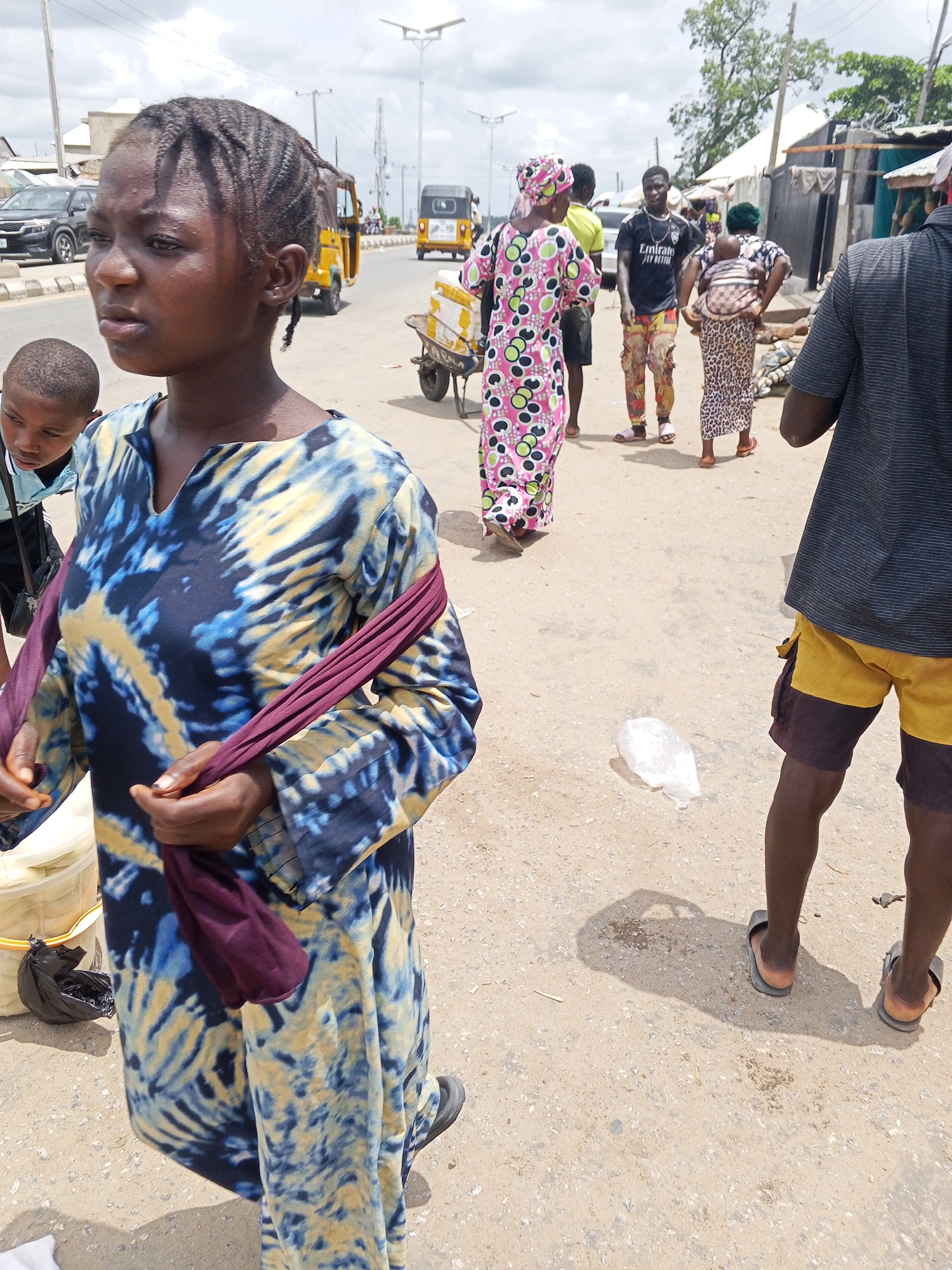 The image shows a street scene in Zango-ndaji, Nigeria. In the foreground, a young woman with braided hair stands wearing a blue and yellow tie-dye patterned dress and a maroon fabric draped over her arm. To her left, a young boy peeks into the frame. In the background, various people are walking and standing on the street. A woman in a long, brightly colored patterned dress walks towards the background, near a yellow tricycle and a man pushing a wheelbarrow. Another man in a black t-shirt and patterned pants is visible, along with a woman carrying a child on her back. A man in shorts and sandals partially obscures the right side of the frame. A plastic bag lies on the ground near him. The setting is a dusty street with buildings and power lines visible in the distance, with cars parked and in motion. The sky is overcast.