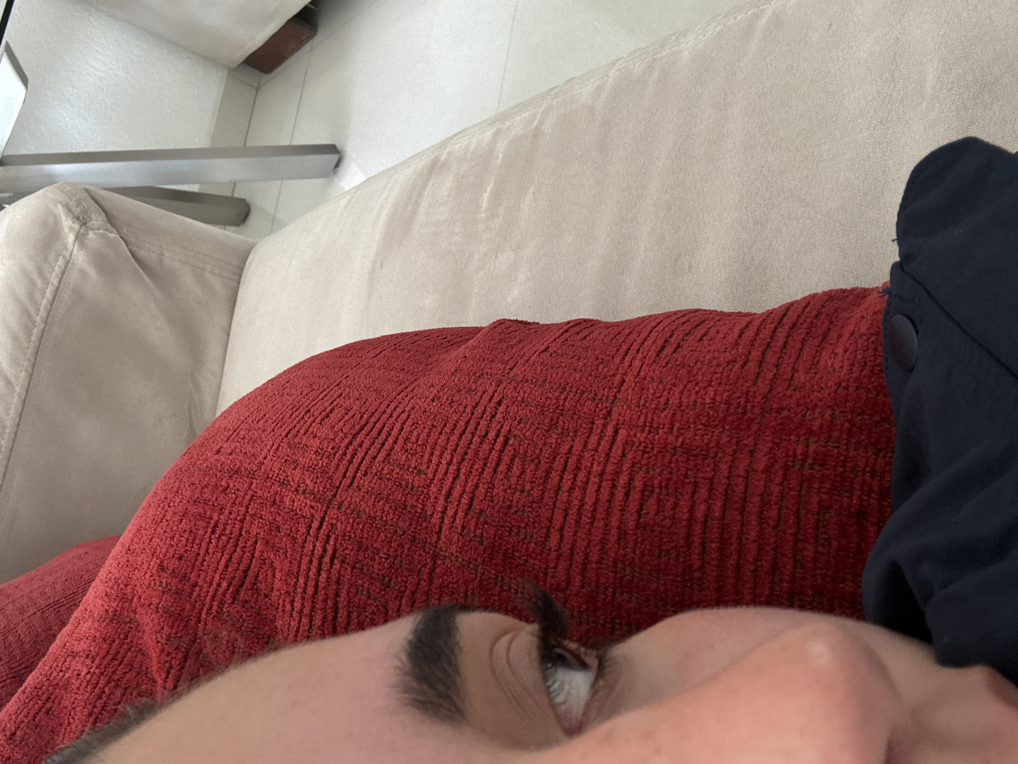 A close-up shot depicts a person lying on a beige couch. The person is positioned on a red textured pillow, with a partial view of their face, including an eye and eyebrow. A portion of a dark blue garment, potentially a jacket or coat, is visible on the right side of the frame. The environment suggests an indoor setting, possibly a living room. The location in Santa Clara Ocoyucan, Mexico, is not deducible from the image.
