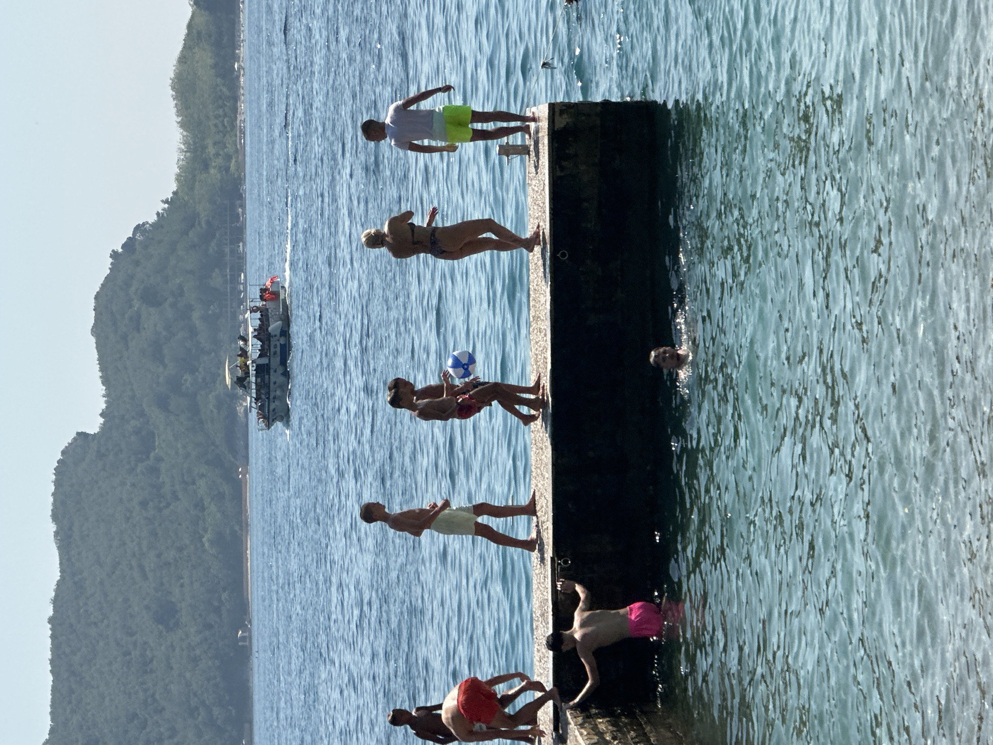 The image depicts a sunny day at a waterfront in Budva, Montenegro. Several individuals, mostly young people, are gathered on a concrete pier and in the clear blue water. On the pier, a group of young men are seen in various states of activity. One boy in red shorts is bending over near the edge, while another, wearing light-colored shorts, walks along the pier. Two other boys are walking together, one holding a colorful beach ball. Further along the pier, a woman in a bikini and a man in a white t-shirt and bright yellow shorts stand facing each other. In the water, a young person in bright pink shorts is partially submerged near the pier's edge, holding onto the side. Another person is swimming in the foreground. In the background, a pleasure boat with passengers is cruising on the sea, with a lush, green, mountainous coastline rising behind it. The water is calm with gentle ripples, and the sky is clear.