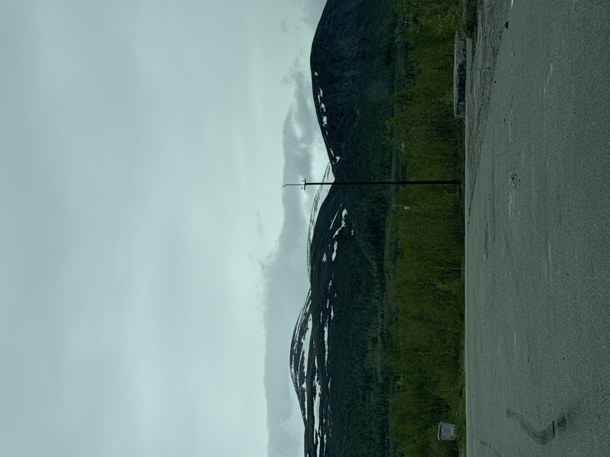 The image depicts a mountainous landscape adjacent to an asphalt road. The sky is overcast. A large, dark mountain with patches of snow covers the majority of the left side. The mountain is next to a road. A pole is visible between the mountains and the sky. The location is suggested to be in the city of Kirovsk, Russia.
