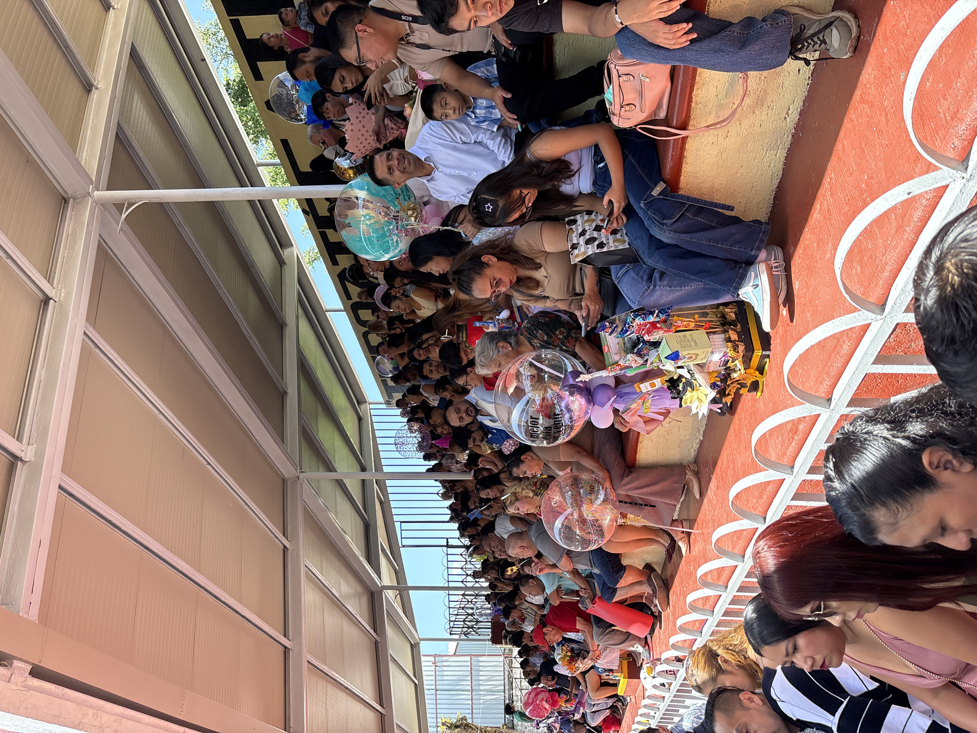 An outdoor tiered seating area, likely a stadium or event venue, is filled with spectators. The seating is a vibrant red, with white railings separating the rows. A yellow retaining wall runs behind the seats. Above, a white roof structure supported by white beams provides shade. The background reveals trees and a clear blue sky.
In the foreground, several people are seated, some looking at their phones, others engaged with the event. Several balloons are visible, including clear, round ones with confetti inside, and a large teal balloon with a printed image. Some attendees hold bouquets of flowers. The people are dressed in casual summer attire, suggesting warm weather. The scene appears to be from a graduation or similar celebratory event, given the balloons and flowers.
The location is specified as Tejalpa, Mexico, suggesting a community gathering in that region. The overall atmosphere is one of celebration and community engagement, with people of various ages gathered to watch an event.