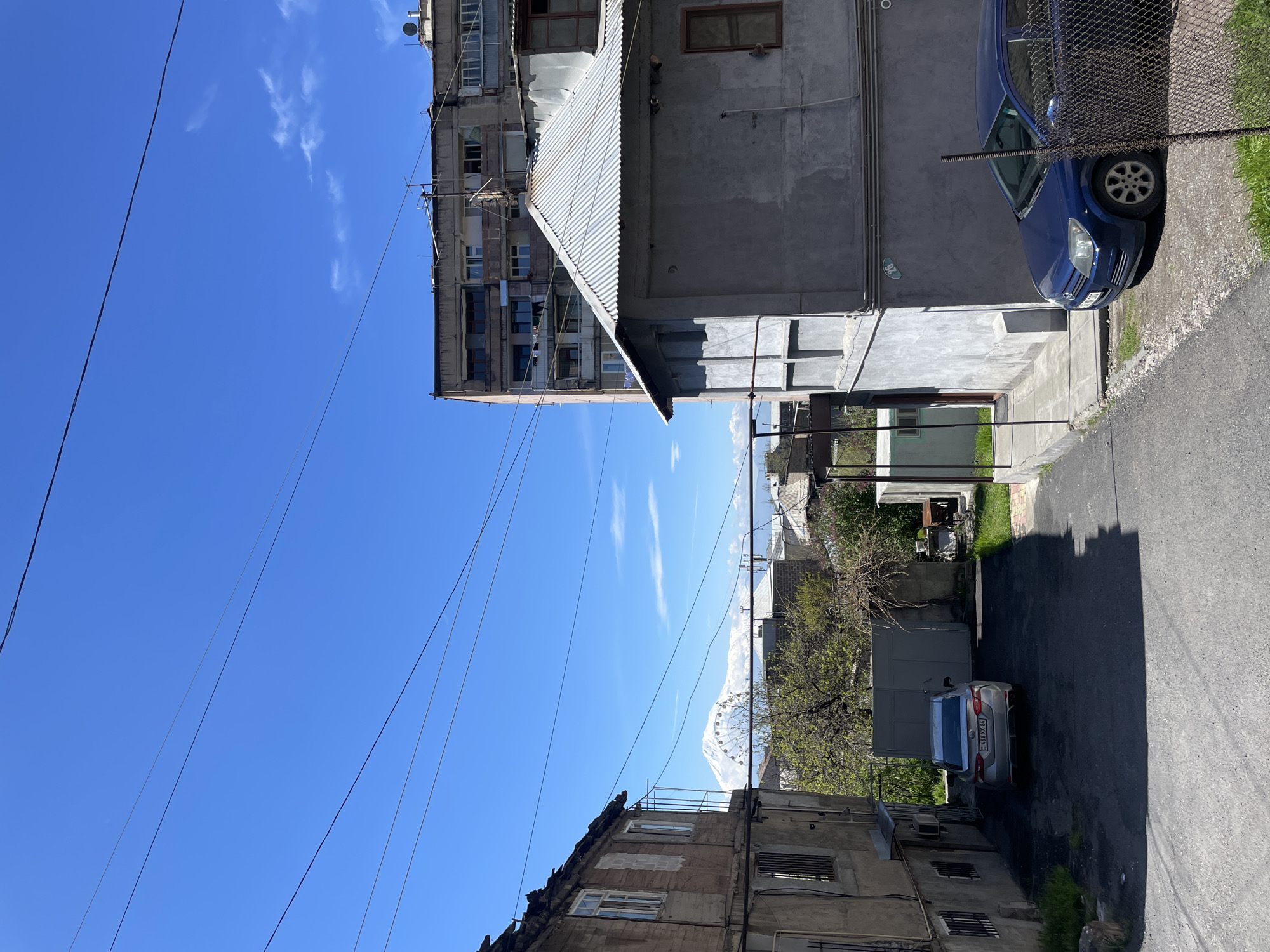 The image presents a sunlit urban alleyway or street scene in Yerevan, Armenia. A vibrant, clear blue sky with sparse, wispy white clouds stretches across the top, intersected by numerous dark utility wires.
In the distant background, a majestic snow-capped mountain range, characteristic of Mount Ararat, rises prominently. Below it, a large white Ferris wheel is partially visible, nestled among buildings and trees.
On the left side of the frame, a multi-story apartment building with a weathered concrete facade, featuring multiple windows and balconies, stands. Further down this side, another building shows a mix of brick and concrete.
On the right, closer to the viewer, a concrete building with a distinctive corrugated metal roof extension is visible. A blue hatchback car is parked against its wall, partially obscured by a chain-link fence. A small, circular sign, possibly reading "92," is affixed to the wall near this car. Further down the paved alley, a silver sedan is parked in front of a gate, displaying the license plate "48 XX 64".
The paved ground of the alley shows distinct shadows, indicating midday or early afternoon under bright, clear weather conditions. Patches of green grass and trees, some with emerging leaves, dot the edges of the path. The scene is static, depicting an ordinary day in a residential area with typical post-Soviet architectural elements.
