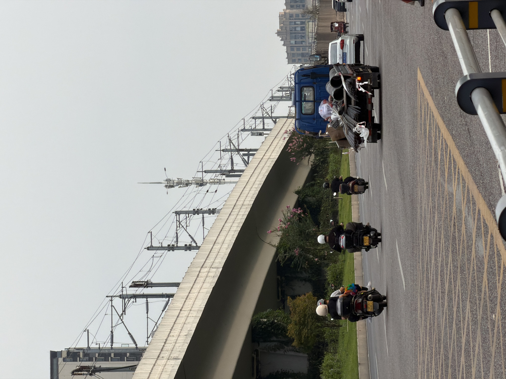 The image depicts a street scene in Jiangjin, China, characterized by an elevated road structure overhead. The road is paved with asphalt and marked with yellow and white lane dividers. In the foreground, several motorcycles are traveling on the road. The first motorcycle, on the left, has two riders, one wearing a white helmet and the other a black helmet. The second motorcycle, also with two riders, is further ahead and to the right, with both riders wearing black helmets. A third motorcycle is visible on the far right, also with a rider wearing a black helmet.
In the middle ground, a blue truck is driving, carrying a load of pipes and rebar, some of which are wrapped in white plastic. To the right of the truck, a white car is also in motion. Further in the distance, a red three-wheeled vehicle and other vehicles are present. The roadside is lined with green vegetation, including trees and bushes. In the background, buildings and communication towers are visible under a hazy sky. The elevated road structure, likely a viaduct or overpass, is supported by concrete pillars and features overhead power lines.