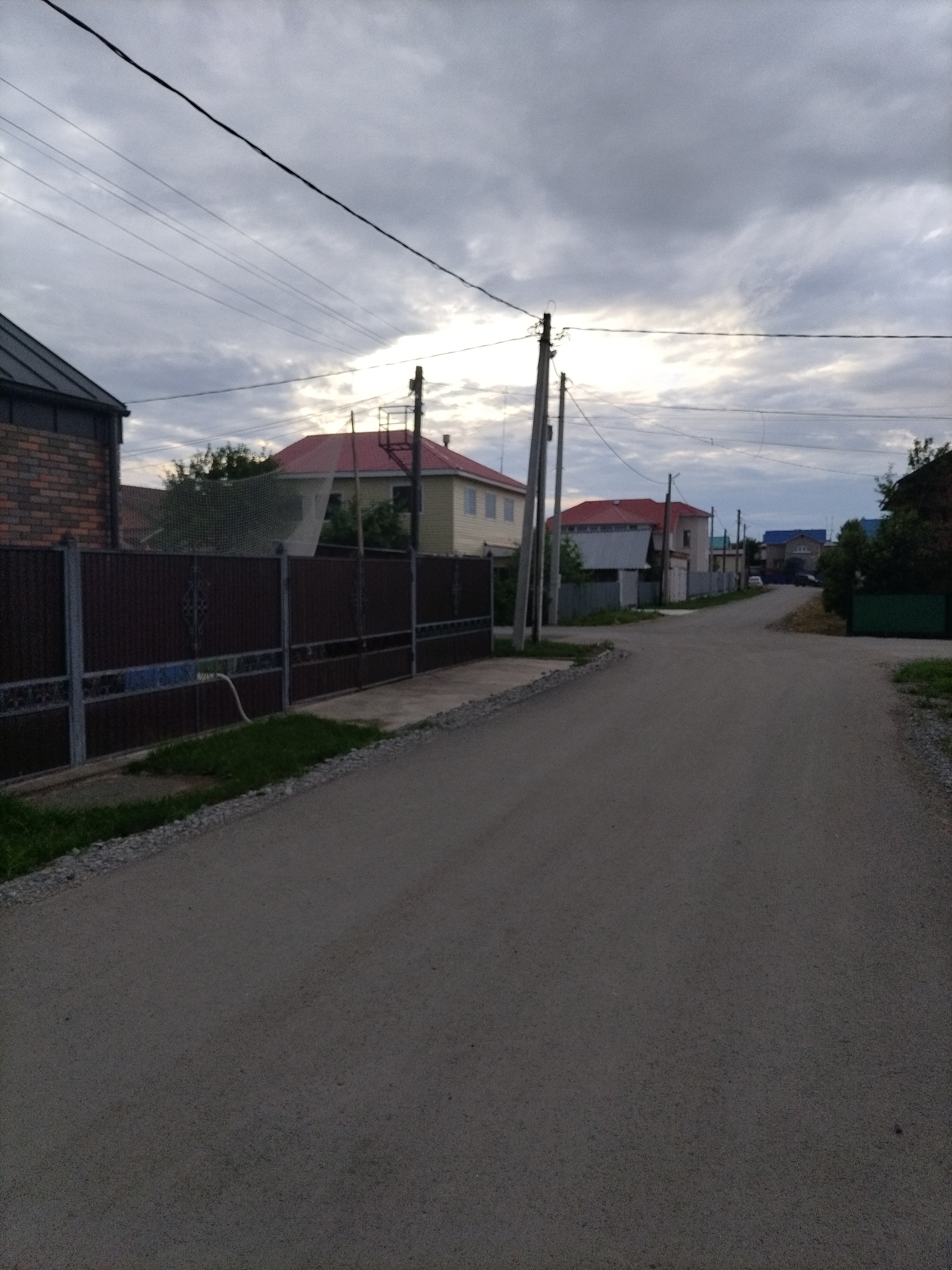 This media file depicts a quiet, rural street scene in the village of Nizhnyaya Makhtama, Russia, under an overcast sky. The road, paved with asphalt, stretches into the distance, curving slightly to the right. On the left side of the road, a tall, dark brown metal fence with decorative elements runs along the property line of a brick building with a dark roof. Beyond the fence, a light-colored house with a red roof is visible, partially obscured by trees and a white net. Further down the street on the left, more houses with red roofs and lighter facades are present.
On the right side of the street, there are also houses visible, one with a dark green gate and another with a blue roof further in the distance. The sky is filled with heavy, gray clouds, suggesting a cloudy or possibly impending rain. The presence of power lines and utility poles indicates that this is a residential area with access to electricity. The scene appears to be a typical day in a Russian village, with no active events taking place. The overall mood is peaceful and somewhat subdued due to the weather. The time of day is likely late afternoon or early evening, given the diffused light. No text is visible in the image.