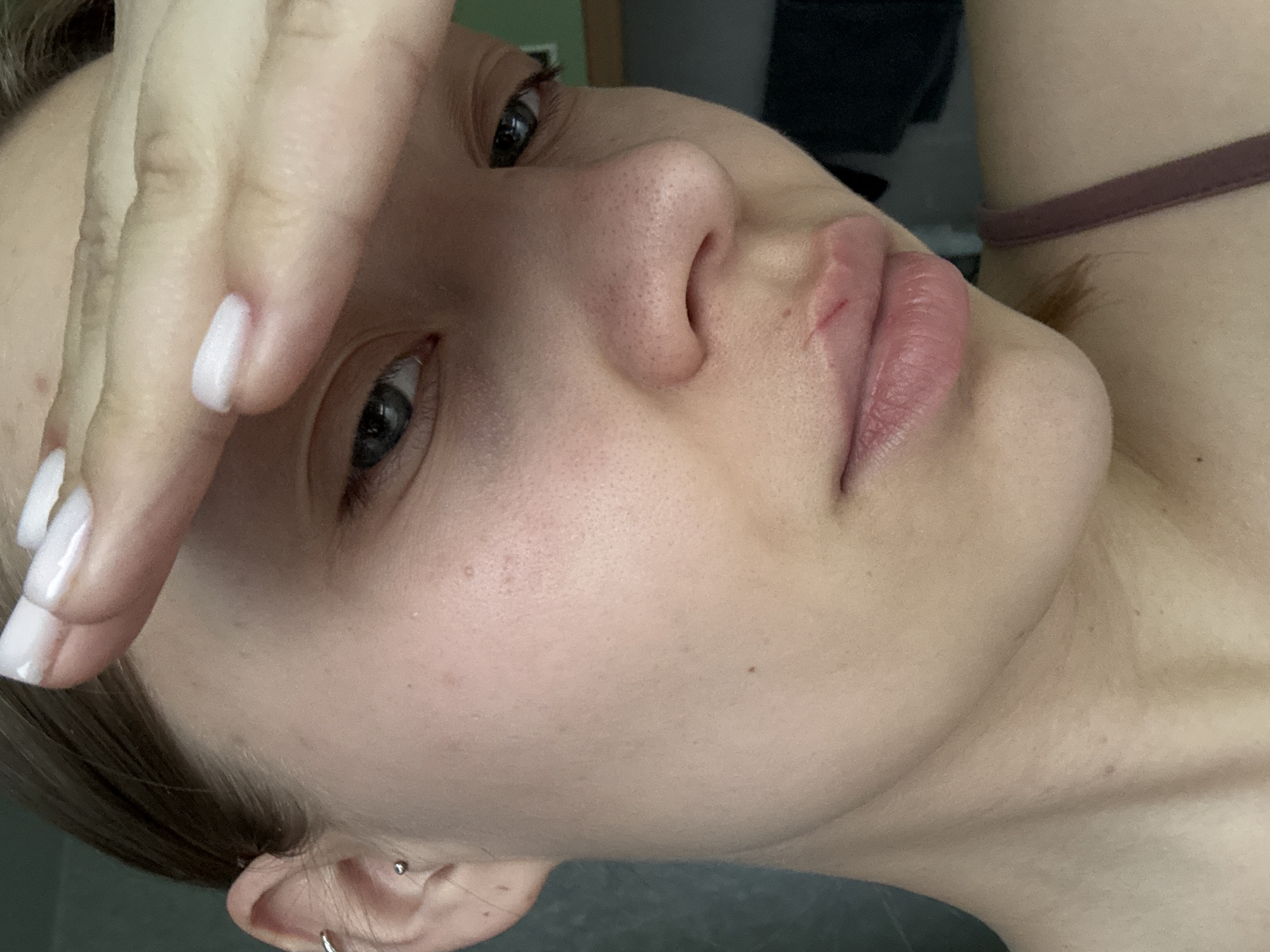 A close-up photograph shows a person's face and part of their hand. The person is fair-skinned with light-colored hair visible in the periphery. Their eyes are open, and they have a small piercing in their ear. The hand is positioned near their forehead, with fingers slightly spread. The fingernails are manicured with a white polish. The background is out of focus, but suggests an indoor setting with indistinct objects. The person is wearing a strap, possibly of a garment, around their neck. No information suggests the location is Chaikovsky, Russia.
