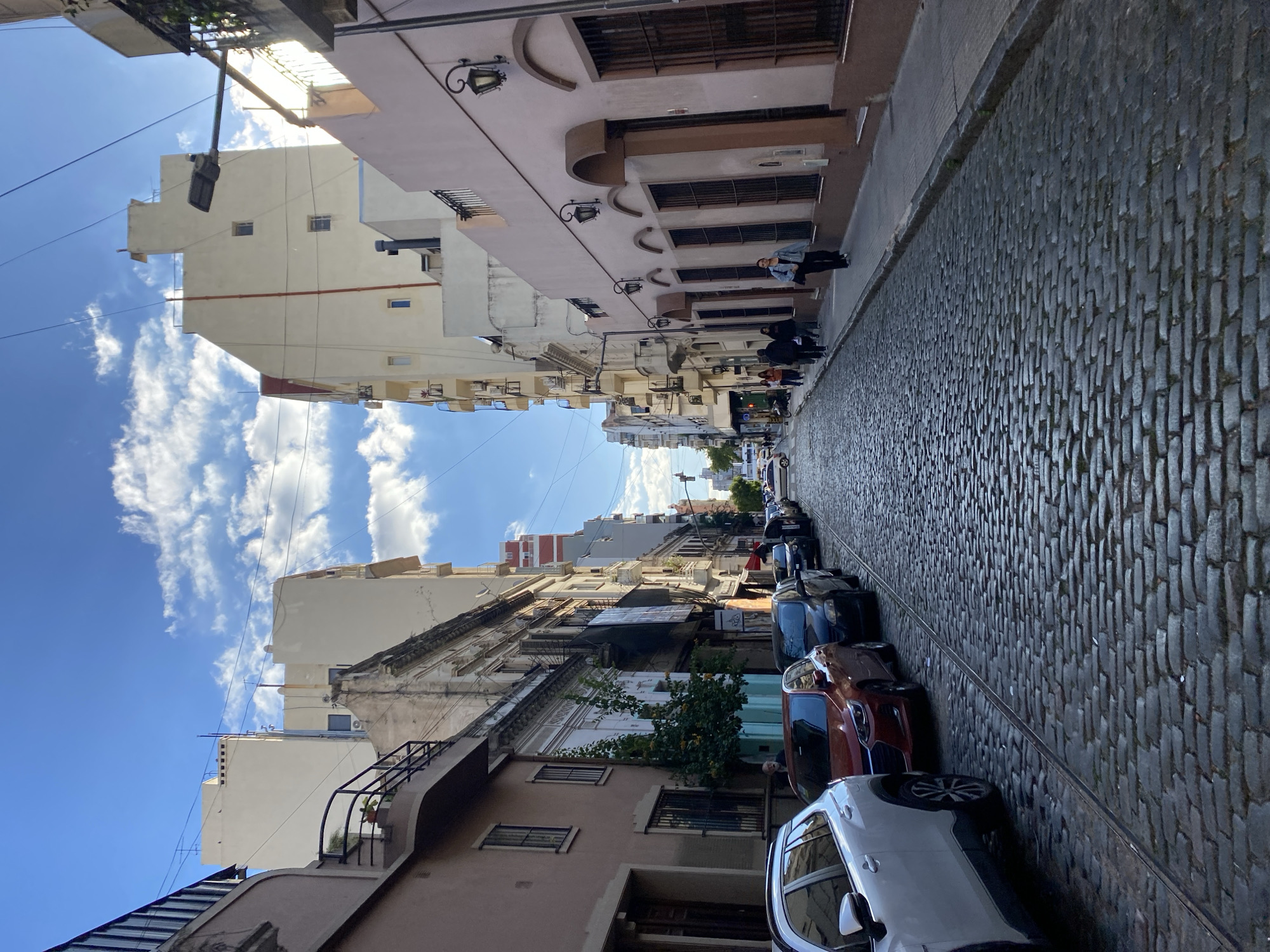 The image depicts a narrow street in Buenos Aires, Argentina, characterized by cobblestone paving. Buildings line both sides of the street, varying in height and architectural style, with facades ranging in color from beige to pink. Some buildings feature wrought-iron balconies and decorative molding. Parked cars occupy the right side of the street. Several people are walking on the sidewalk on the left side. The sky is visible at the end of the street.