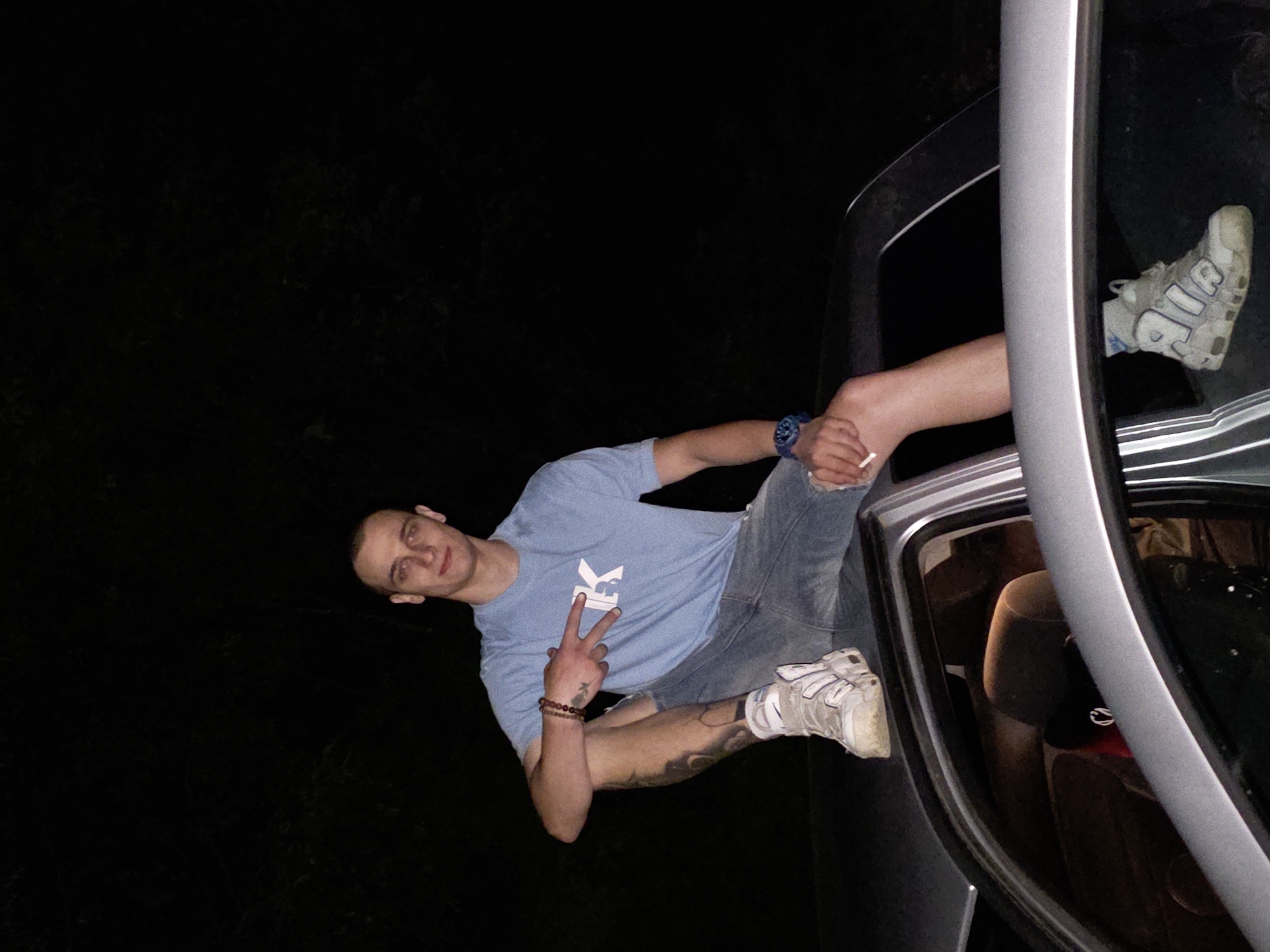 A medium-shot of a man sitting in the open window of a dark gray car. The man is Caucasian, has short, dark hair, and is wearing a light blue t-shirt, denim shorts, and white sneakers. He is holding a cigarette in his left hand and making a peace sign with his right hand. He is wearing a watch and bracelets. Part of a tattoo is visible on his leg. The interior of the car is visible, showing a brown car seat and a red hat. The background is dark, suggesting nighttime. The location is Lyaskovo, Bulgaria.