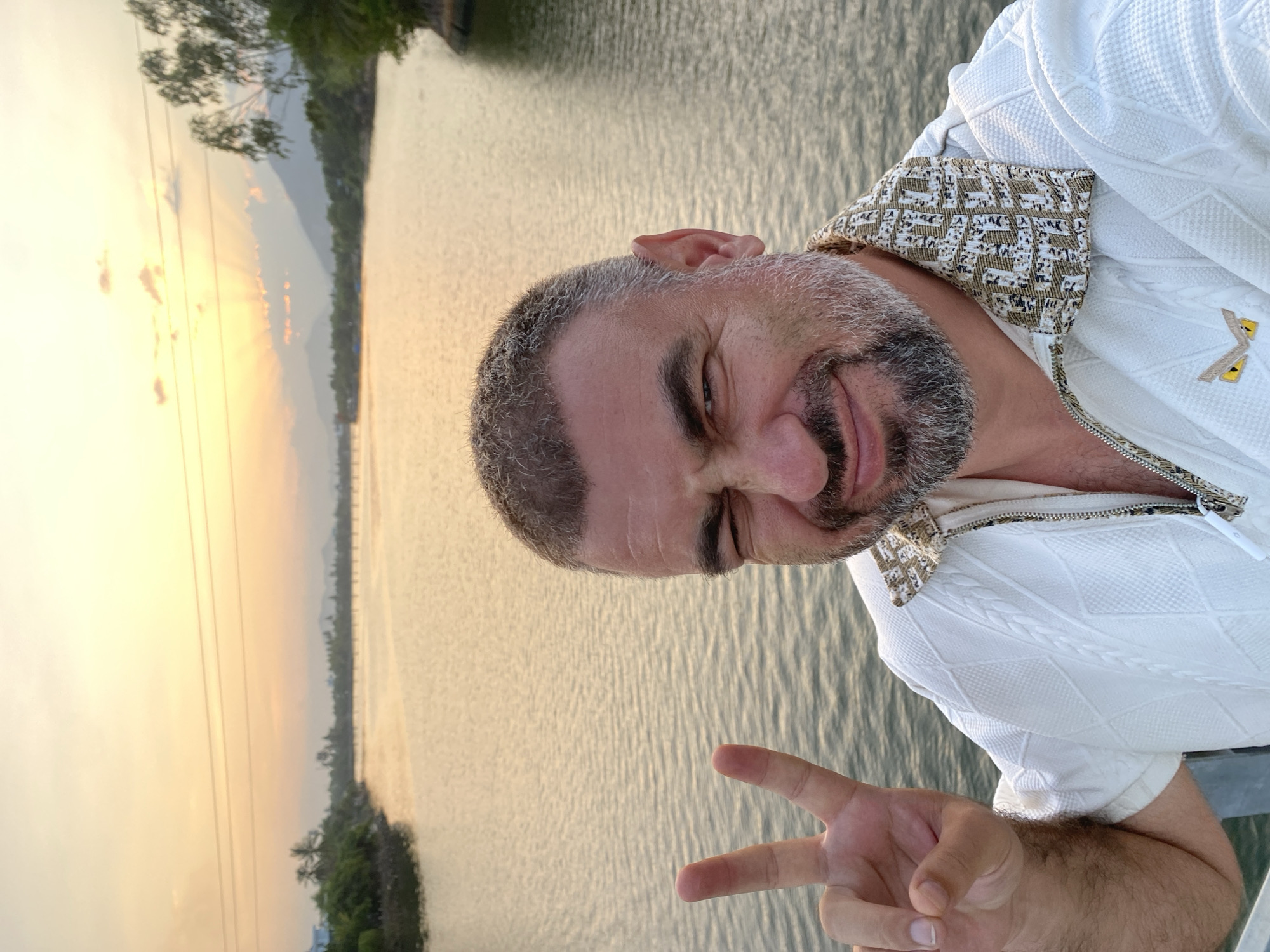 A man with short, grey-flecked hair and a beard winks and makes a peace sign with his left hand in a selfie. He is wearing a white, textured polo shirt with a zippered neckline. The shirt has a patterned collar in shades of beige and brown. A small, yellow, owl-like logo is visible on the left side of his chest.
The man is positioned in the foreground, with a body of water behind him. The water reflects the warm, golden hues of a sunset. In the distance, a bridge spans the water, and a hazy shoreline with trees and mountains is visible. The sky is awash with soft orange and yellow tones, with rays of light breaking through clouds. Power lines are visible in the upper part of the frame. The scene is likely a public walkway or bridge in Nha Trang, Vietnam, given the coastal setting and tropical vegetation suggested by the trees.