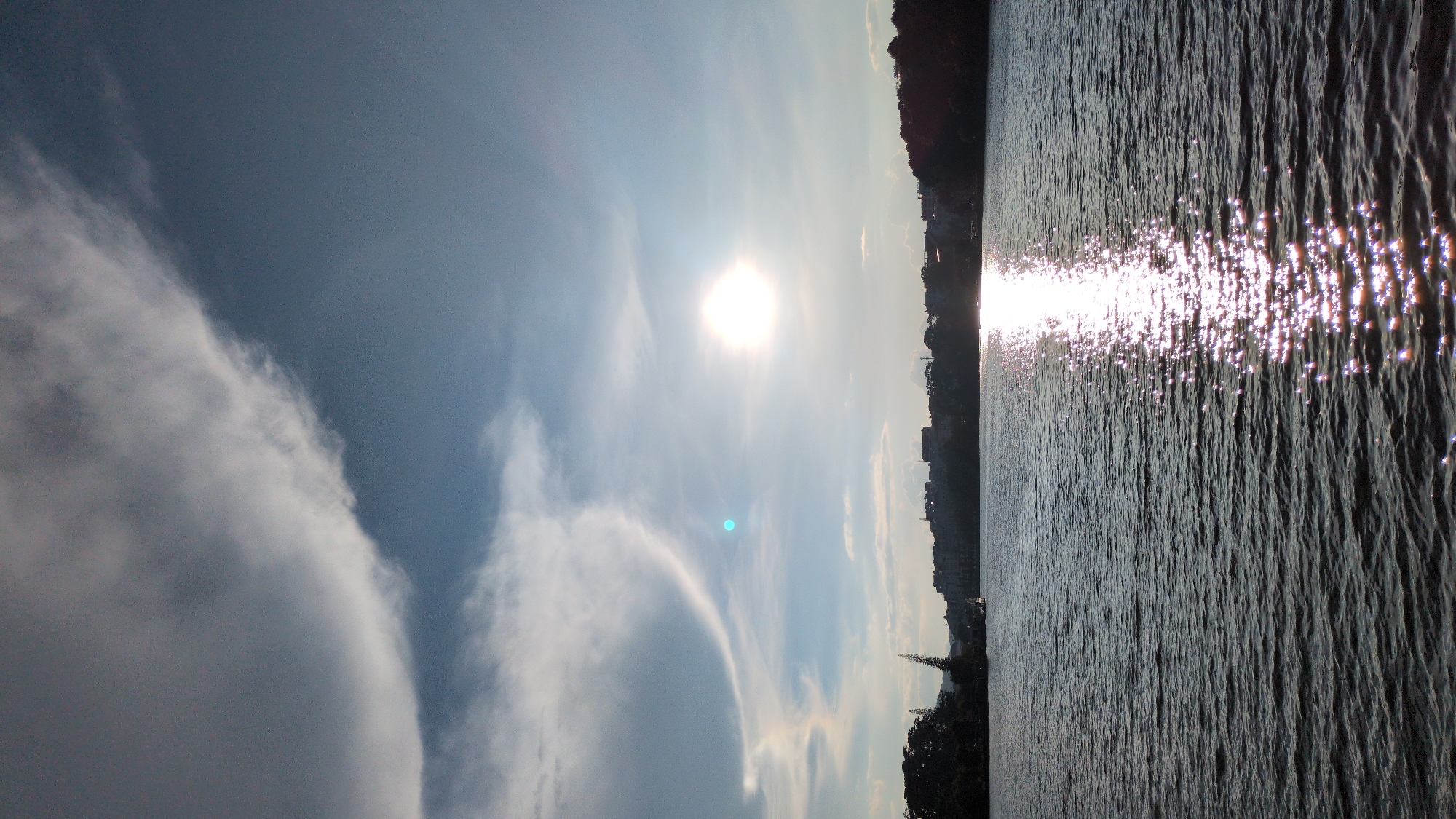 This is a photograph of Xuan Huong Lake in Da Lat, Vietnam, taken from a low angle, likely from a boat on the water. The sun is high in the sky, casting a bright reflection on the water's surface, creating a shimmering path of light. The sky is a pale blue with scattered white clouds. In the distance, a line of buildings and trees forms the shoreline of the city. The water is choppy, with small waves creating a textured surface. The overall scene conveys a sense of calm and natural beauty.