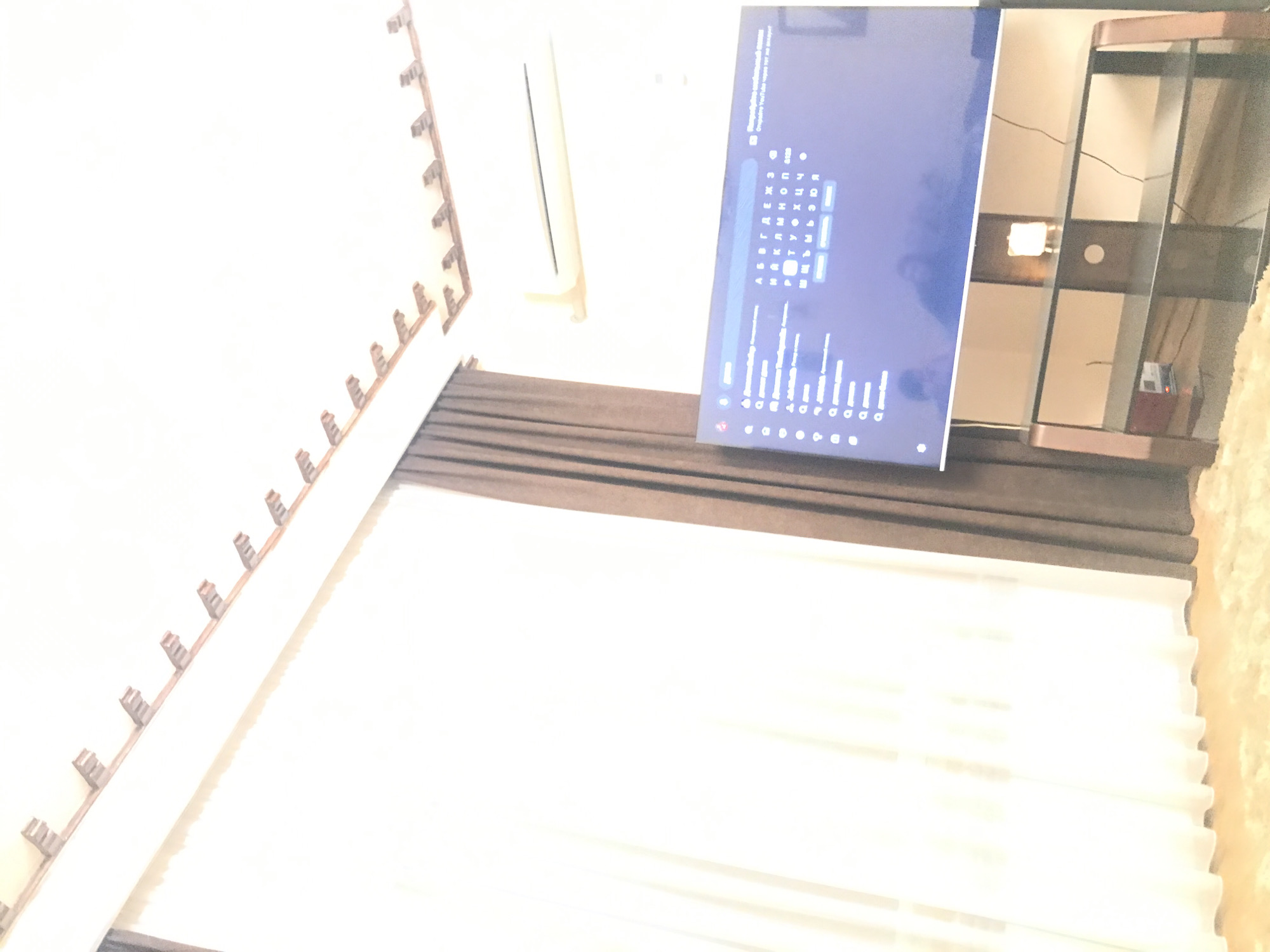 A sideways, blurred photograph shows a room with a TV displaying a screen with Russian text on it, positioned in front of a shelving unit. The wall above the TV has decorative elements along the top edge. Below the TV, there is a curtain and a sheer, white curtain that appears to cover a window. Due to the limited detail and blurred nature of the image, specific features or objects in Yerevan, Armenia, can't be identified.