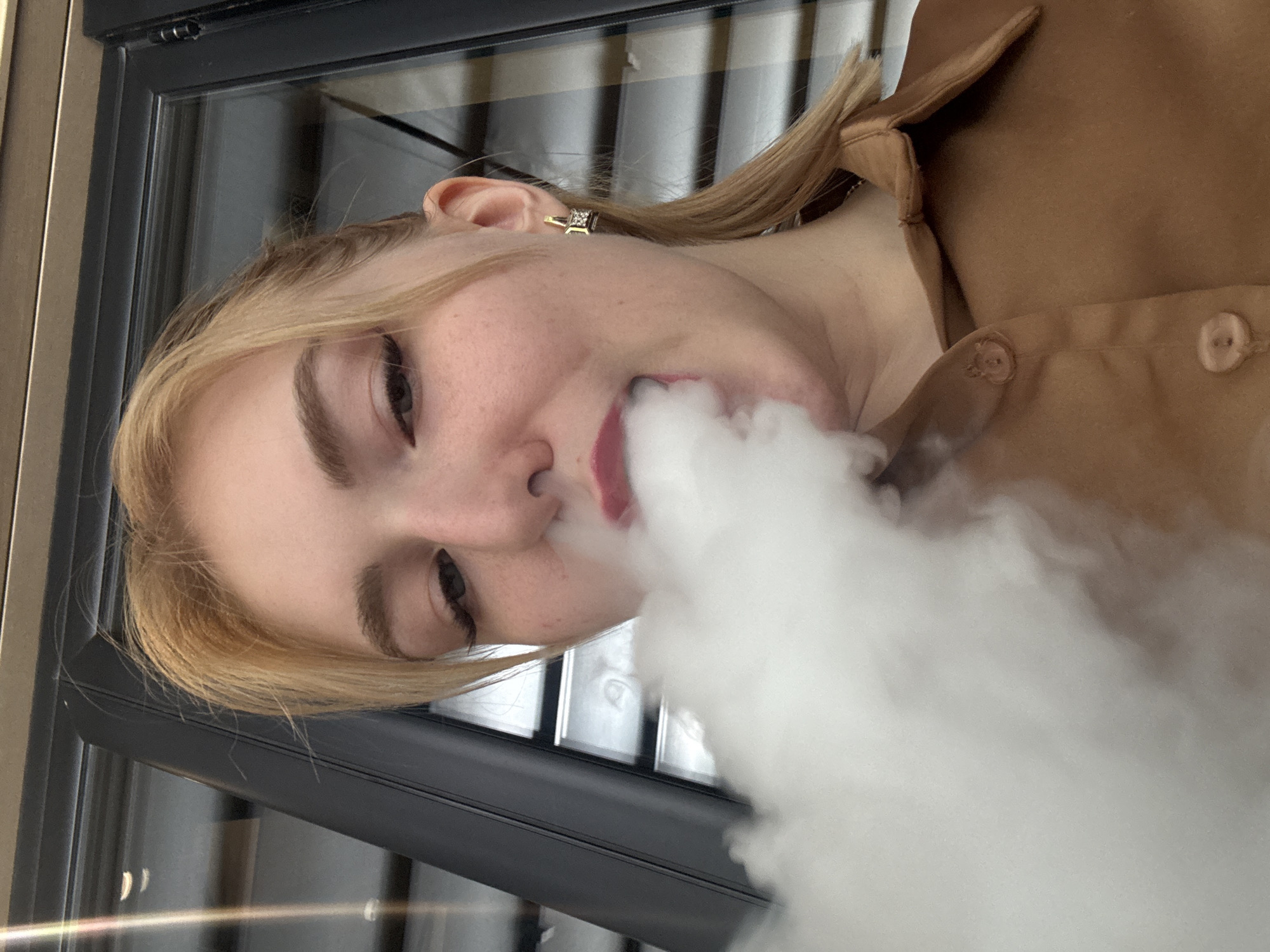 This media file is a close-up, low-angle photograph of a young blonde woman. She is wearing a tan-colored collared shirt with two visible buttons. A large plume of white smoke billows from her mouth, obscuring the lower half of her face and extending outwards. Her left eye is visible, with a light brown iris and dark eyelashes. She has a small, square-shaped earring in her left ear. The background appears to be a window or glass surface reflecting a striped pattern, suggesting an indoor setting. The lighting is moderate, and the overall mood is somewhat obscured by the smoke. No visible text is present in the image. The context provided places the location as Moscow, Russia.
