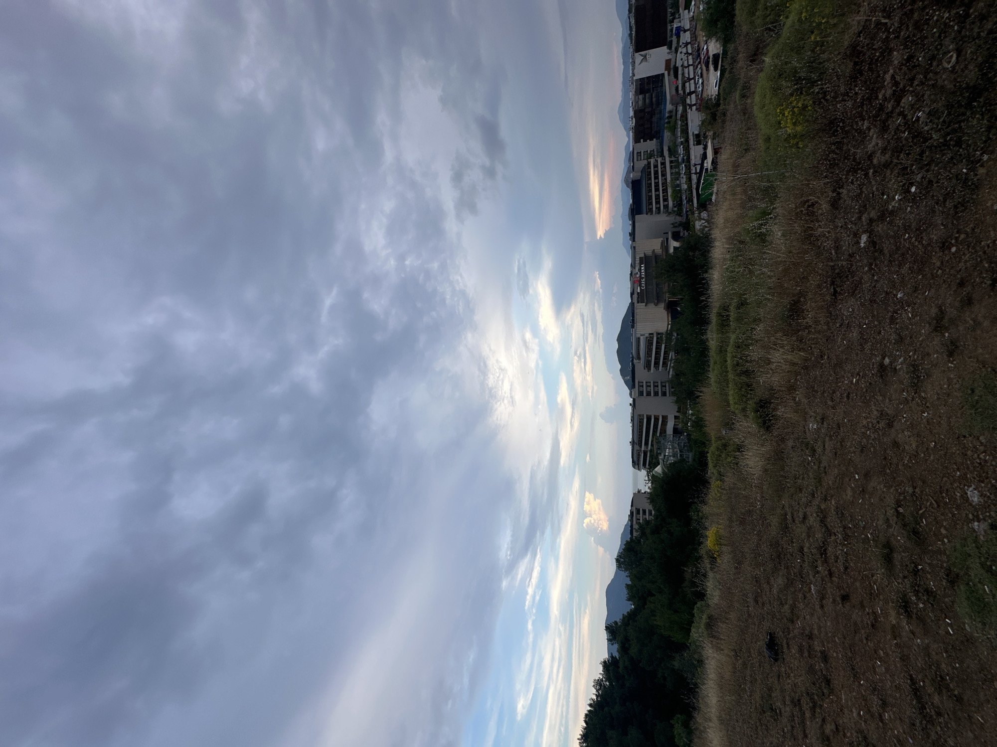 The image depicts a landscape in Fethiye, Turkey, taken from an elevated position. The left side of the frame is dominated by a cloudy sky, with hues of blue and grey. The right side transitions into a view of the city, with buildings visible amongst trees and mountains in the background. In the foreground, there is an area covered with dry grass and vegetation. The perspective is slightly tilted.
