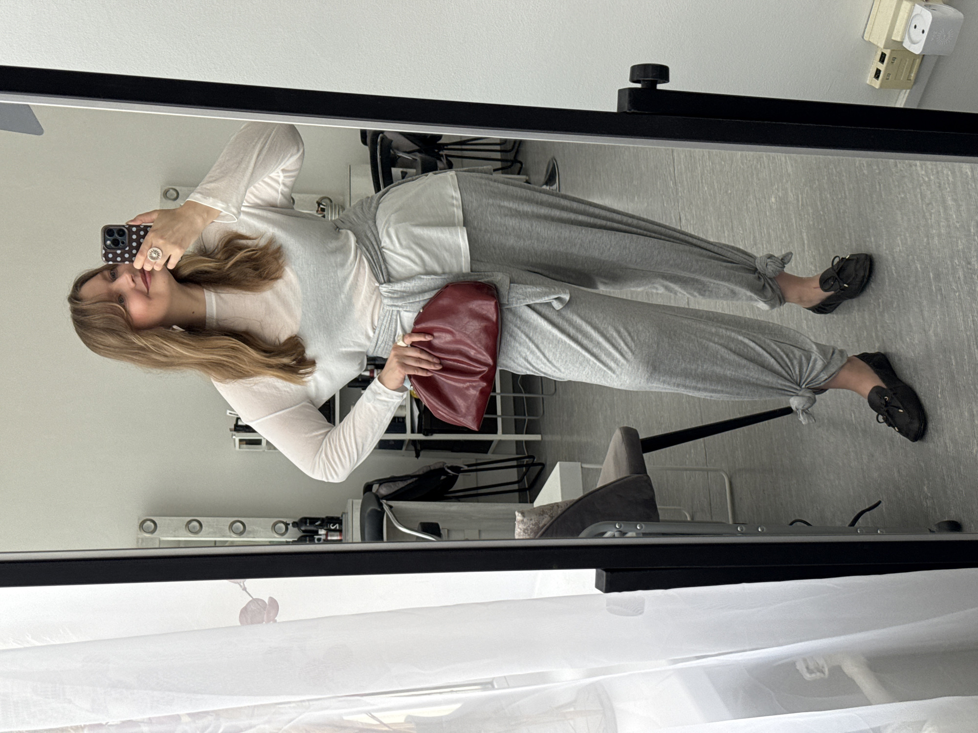 A young woman takes a mirror selfie in an indoor setting. She has light brown, wavy hair that falls over her shoulders and wears a faint smile. She is dressed in a light-colored, possibly sheer, long-sleeved top, under which a white layer is visible. Her lower body is clad in light grey jogger-style pants, which are tied at the ankles, and a matching grey sash or fabric element is tied around her waist.
In her left hand, she holds a dark red/burgundy clutch bag. Her right hand, adorned with a ring featuring a circular design (possibly a sun or flower), holds a smartphone with a dark brown case patterned with white polka dots, which she is using to capture the image. White earbuds are visible in her ears. She completes her outfit with black flat shoes or loafers, featuring a small bow detail.
The background, reflected in the mirror, reveals light-colored walls and a light grey floor. Various elements of the room are subtly visible, including what appears to be a power outlet or adapter mounted on the wall, parts of a chair with a grey back and silver legs, and a rack or shelving unit. A white sheer curtain is reflected at the bottom of the frame. No discernible text is visible on clothing or accessories, apart from the pattern on the phone case.