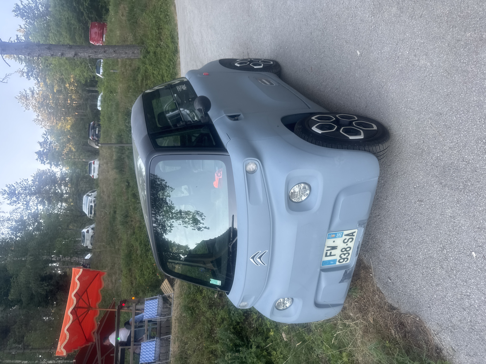 In Maisod, France, a light blue Citroën Ami electric car is parked on a paved surface next to a grassy slope. The car has a black roof and unique black and white wheel covers. Its license plate reads "FW-938-SA". Behind the car, on the slope, several other vehicles are parked, including a white SUV, a black SUV, and a red van. A tall, thin tree stands to the right of the car. In the background, dense green trees and foliage are visible, with a bright sky overhead. To the left of the car, a red umbrella shades a seating area with checkered blue and white chairs, where a person in a white shirt is seated.