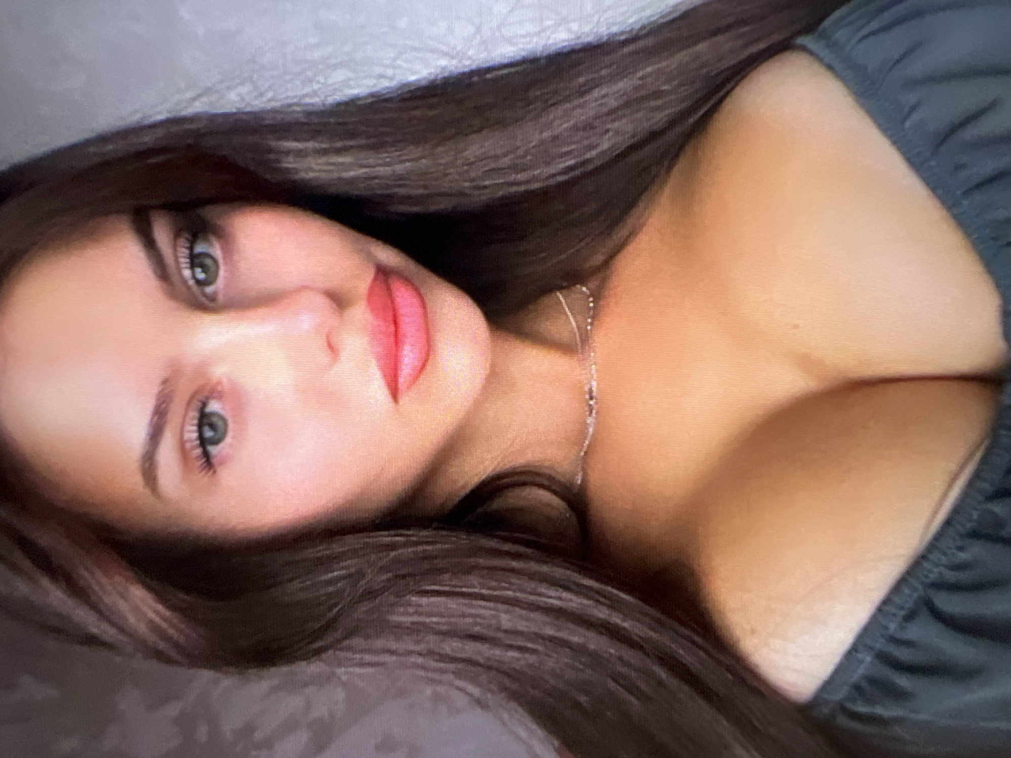 The image is a close-up portrait of a woman with long, dark hair framing her face. She has green eyes and full, pink lips. She is wearing a dark, off-the-shoulder top and a delicate silver necklace. The background is out of focus, appearing to be a textured wall. The lighting is soft, highlighting her facial features and décolletage. The scene is indoors, with no discernible city or geographical information.