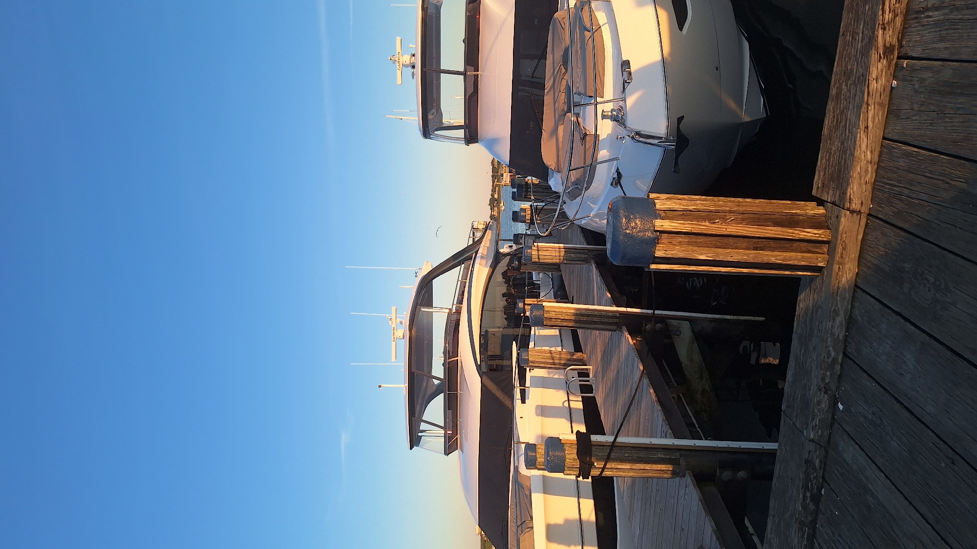 Two large, white yachts are docked side-by-side at a wooden pier under a clear, blue sky. The yacht on the left is larger and appears to be a sport yacht with a sleek, modern design and a dark tinted canopy. The yacht on the right is also a luxury yacht, with a white hull and a covered foredeck. The pier is constructed of weathered wooden planks and features several wooden posts with blue caps. The scene is illuminated by the warm, golden light of a sunset or sunrise, casting long shadows. In the distance, other boats and a shoreline with buildings can be seen. The overall impression is one of a peaceful and affluent maritime setting.