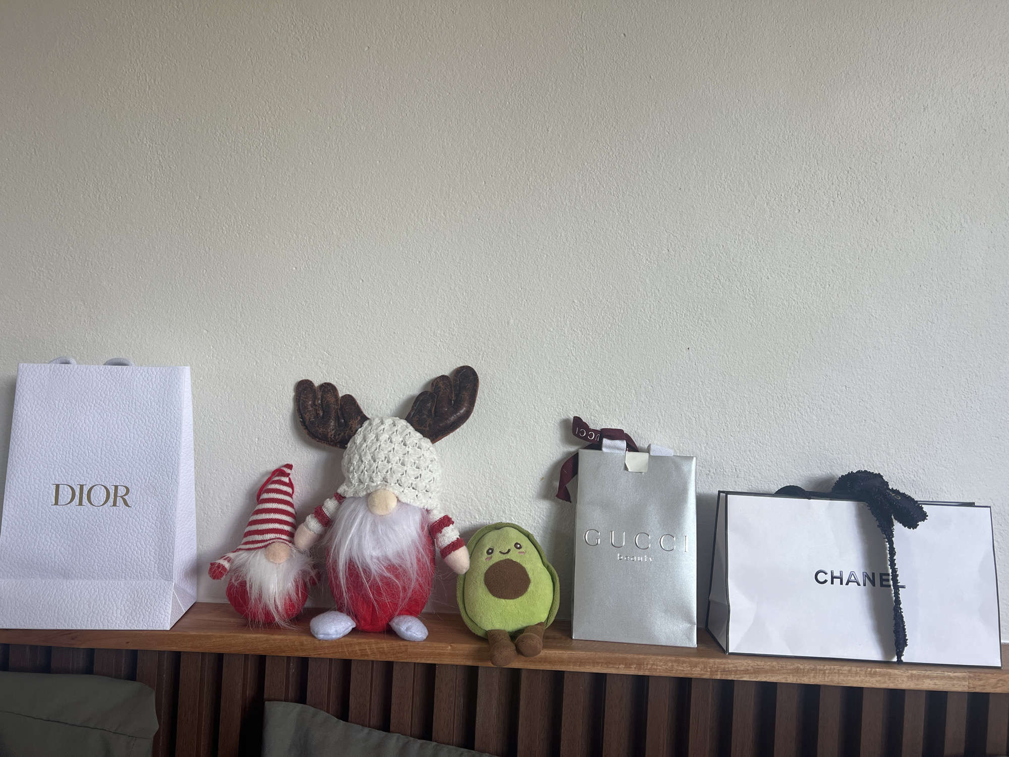 On a wooden shelf against a light beige wall in what could be a residence in บ้านบางรักษ์, Thailand (though this cannot be definitively confirmed from the image alone), there are four shopping bags and three small plush toys. From left to right, there is a white "DIOR" shopping bag, a small gnome with a red and white striped hat, a larger gnome with brown antlers and a white crocheted hat, a small avocado plush toy, a silver "GUCCI" shopping bag, and a white "CHANEL" shopping bag with a black ribbon hanging out. The focus is on the arrangement of the objects on the shelf.