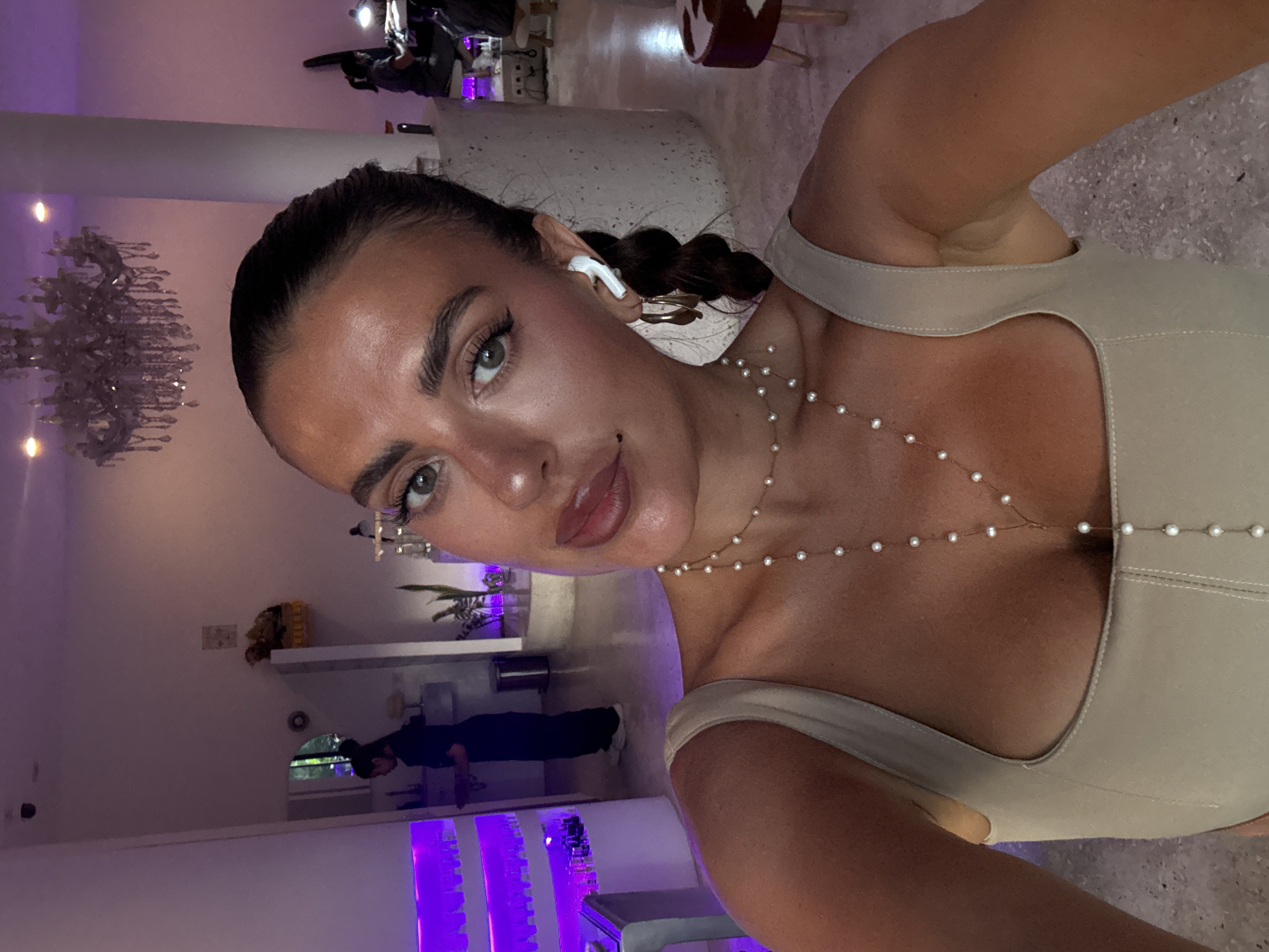A light-skinned woman with dark hair braided in a single braid is taking a selfie in what appears to be an indoor business establishment, potentially a salon or spa. She is wearing a beige top with a low neckline and a delicate pearl necklace. An earpiece is visible in her left ear. Behind her, the scene is illuminated by purple lighting fixtures, giving the space a modern, upscale feel. A person is visible in the background. A chandelier is visible in the distance, and other decorative elements such as plants and displayed products suggest a stylish environment. Considering the location is North Kuta, Indonesia, this scene is likely in a high-end business catering to a sophisticated clientele.
