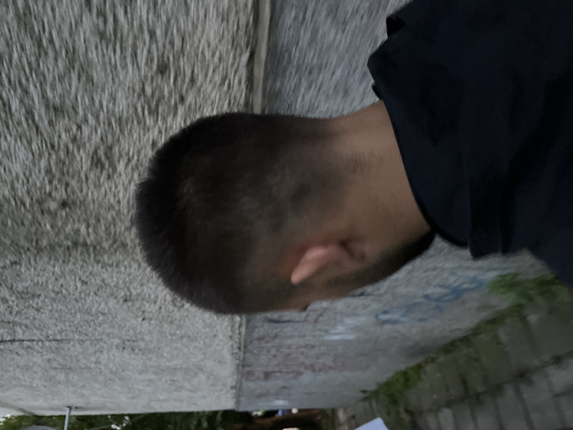 This media file captures a close-up, profile view of a person from the back-left side, appearing to look downwards or to their left. The person has a very short, buzz-cut hairstyle and is wearing a dark-colored top, possibly black or navy. Only the back of their head, a visible ear, and a portion of their shoulder are discernible.
The setting appears to be an outdoor urban or semi-urban environment in Shumen, Bulgaria. To the person's left is a prominent, rough-textured grey concrete or stone wall, which dominates the left side of the frame. Below the person and extending towards the right, the ground surface is visible. This surface features faint blue graffiti, with some visible numbers like "25" and other illegible script or symbols. Patches of green vegetation are also present near the ground. Further into the background, what looks like another segment of a concrete structure or wall, possibly part of an underpass or bridge, is seen, with hints of outdoor light and distant greenery.
The lighting is diffused, suggesting either an overcast day or a shaded area, possibly within or adjacent to a concrete structure. No specific activity is taking place; the person is stationary. The original image is rotated 90 degrees clockwise.