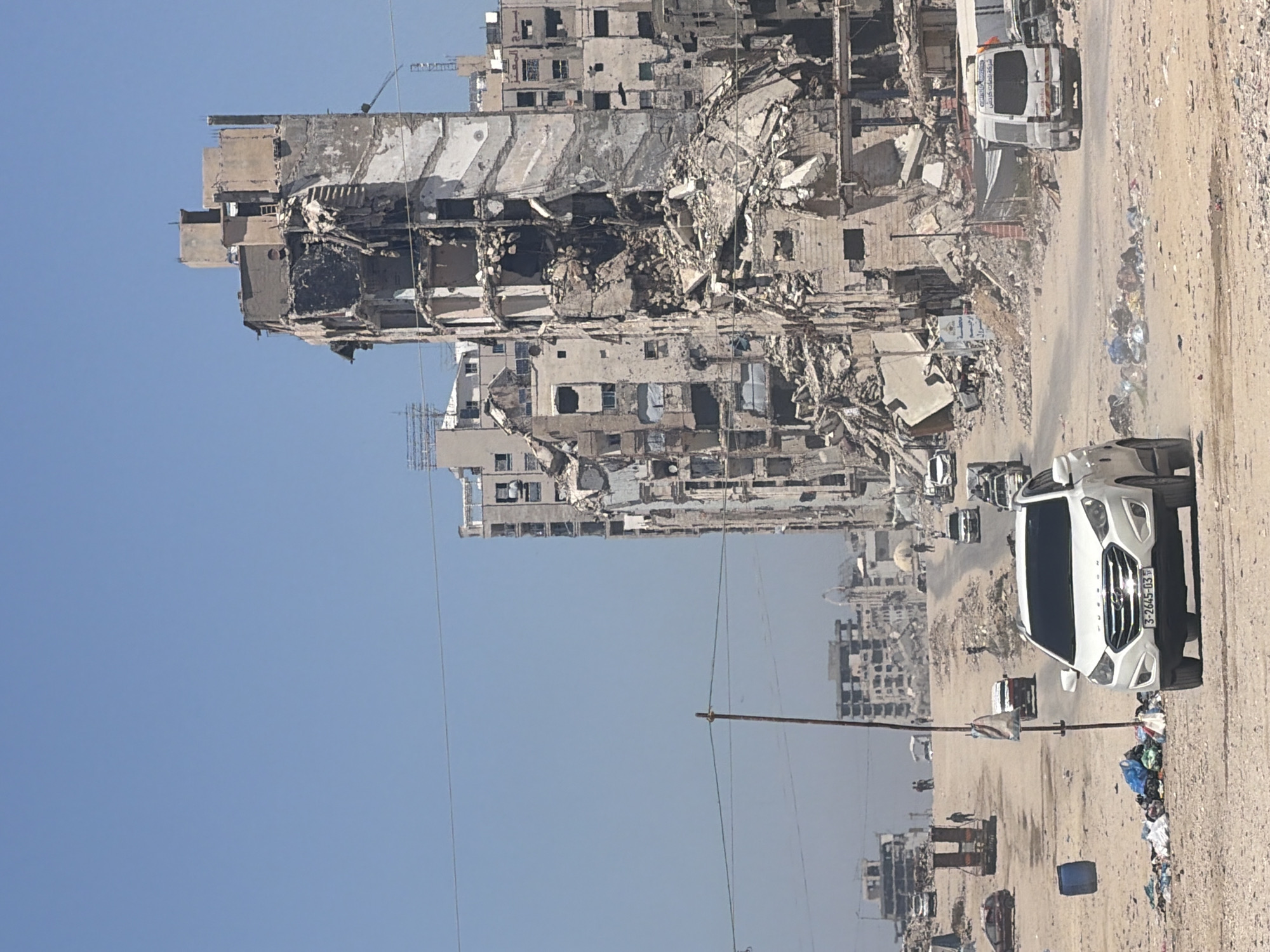 In the city of Unknown, Unknown, a clear, blue sky hovers over a scene of widespread destruction. The foreground is dominated by a dirt road, littered with debris and trash. A white SUV is parked on the right, its license plate reading "3-2645-03". Further down the road, a white van with text on its side is visible, along with several other vehicles.
The background reveals a cityscape ravaged by conflict. Tall buildings stand in various states of ruin, with some completely destroyed, exposing their internal structures. Rubble and debris are scattered across the ground, suggesting a recent catastrophic event. A few people are seen walking in the distance, indicating signs of life amidst the devastation. A utility pole stands on the left side of the road, draped with a bag, adding to the somber atmosphere of the scene.