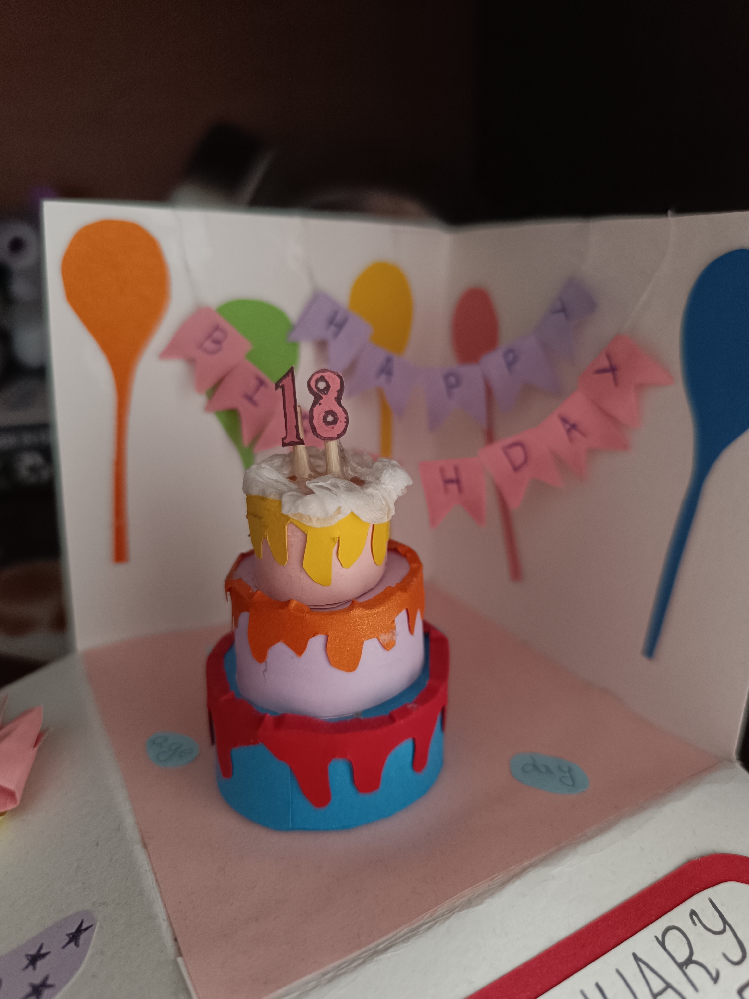 The image shows a handmade birthday card featuring a three-tiered cake with candles indicating the number 18. The cake is decorated with colorful drips, and the card itself has balloon and pennant decorations with the text "HAPPY BIRTHDAY". The background of the card is white. The objects present on the card include the cake, balloons, pennants, and written words. The card appears to be in a closed, open position revealing the inside design. The location is not discernible.