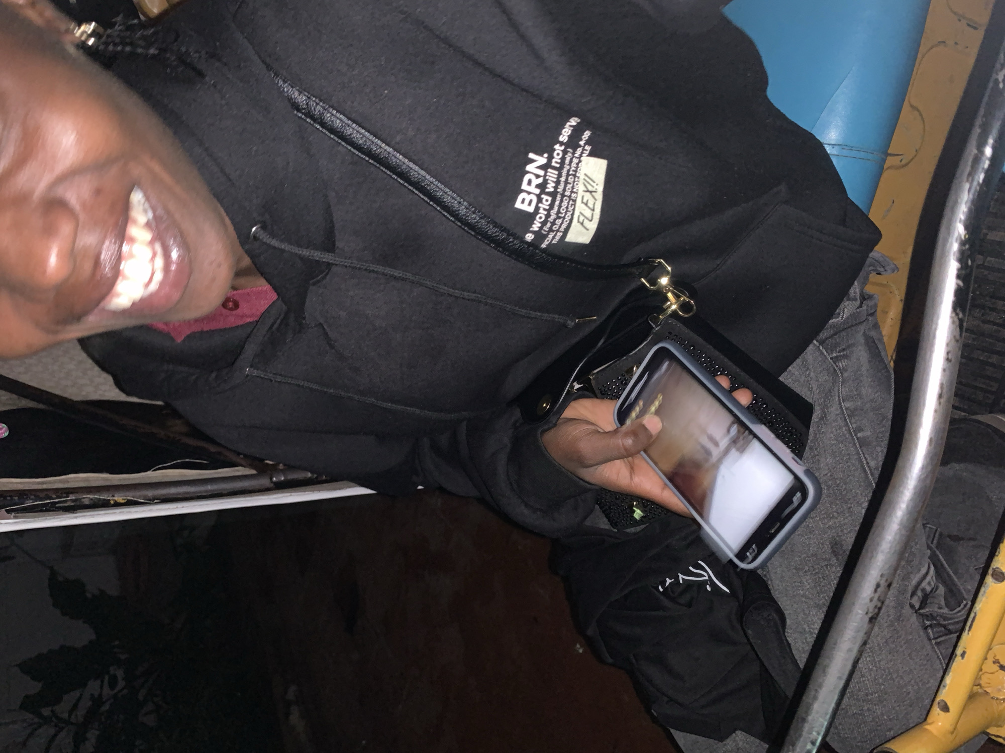 The image shows a person holding a smartphone. The person's face is partially visible, with their mouth open in a smile, revealing their teeth. They are wearing a black jacket with text printed on it. The text reads "BRN.", "e world will not serv", "(For Influencer Marketing only)", "-FICIAL OG. LOGO SOLID TYPE No.A00", and "THIS PRODUCT IS NOT FOR SALE FLEX!!". The person is in what appears to be an indoor setting with some kind of seating that is blue and yellow. The lighting suggests it could be evening or a dimly lit indoor space. The provided location context is Jos, Nigeria, but there are no specific visual cues in the image to confirm this.