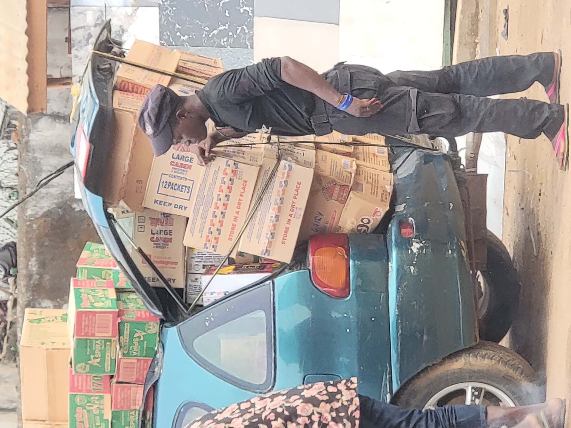 In Maiburiji, Nigeria, a blue/green compact car is seen heavily laden with cardboard boxes, stacked both inside the open hatchback and on the roof. The boxes appear to contain goods for sale or distribution; some are labeled "OXFOR" and "LARGE CABIN CAREN," indicating they hold 12 packets, with instructions to "STORE IN A DRY PLACE." Other boxes labeled "ASPIRA" are also visible.
A man wearing a black shirt, black pants, a baseball cap, and blue wristband is bending over, seemingly adjusting or checking the boxes inside the car. A person wearing a floral patterned dress is partially visible in the foreground, near the car's rear tire. The car is parked outdoors in an urban or semi-urban setting. The background shows a mix of buildings and ground covered with dirt.