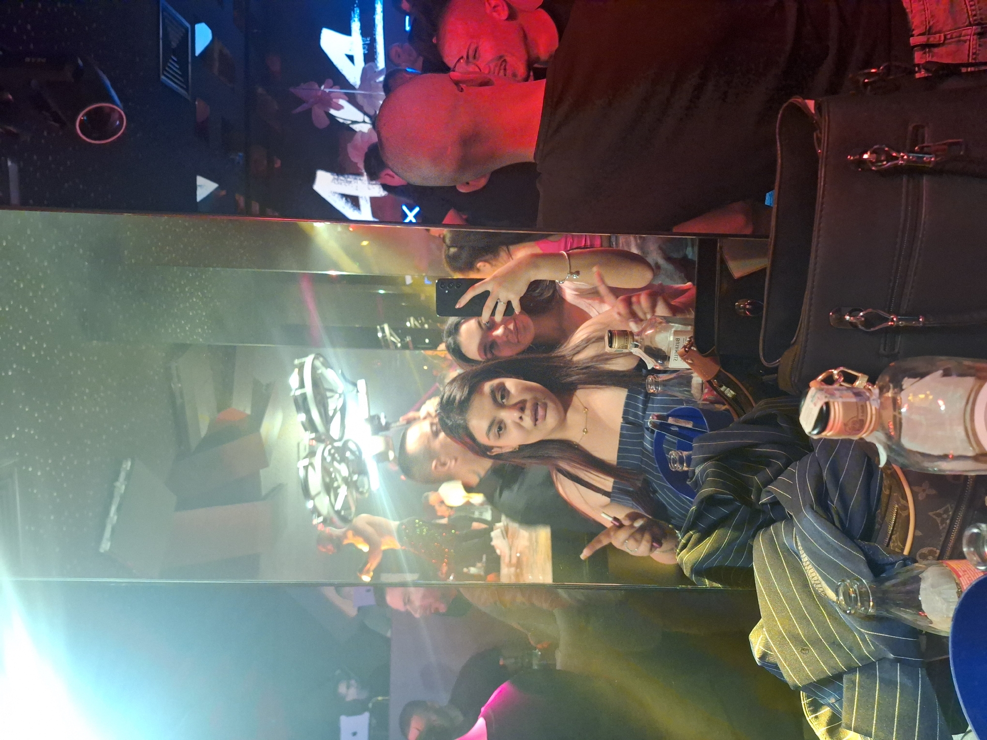 This image is oriented sideways, depicting a lively indoor scene, likely a nightclub or bar in Sofia, Bulgaria, characterized by strong reflections and vibrant artificial lighting.
In the foreground, a young woman with long dark hair, wearing a striped top, looks directly at the camera with a calm expression. She is seated at a reflective surface, likely a table or bar, which horizontally bisects the frame. On this surface, a black structured handbag with prominent metal clasps is visible on the right, next to a draped piece of striped fabric. Several glass bottles, including one that appears to be an alcoholic spirit, and a blue cup are also present. The label of one bottle shows "Bozhilka" or similar text in reflection.
The reflective surface shows reflections of other individuals and the venue's interior. A young woman with a smartphone is seen, smiling and seemingly taking a selfie or video. Another person's face is partially visible beside her. In the background, or reflected above, a man with a shaved head and glasses looks away, his profile illuminated by a strong red light. Behind him, glowing light installations on a dark wall display a large white number "4" and a stylized "X".
The overall setting suggests a nighttime social gathering, with bright, colored artificial lights (red, blue, yellow) creating a dynamic and somewhat obscured atmosphere. The presence of drinks and the pose of the individuals indicate a social event or party.