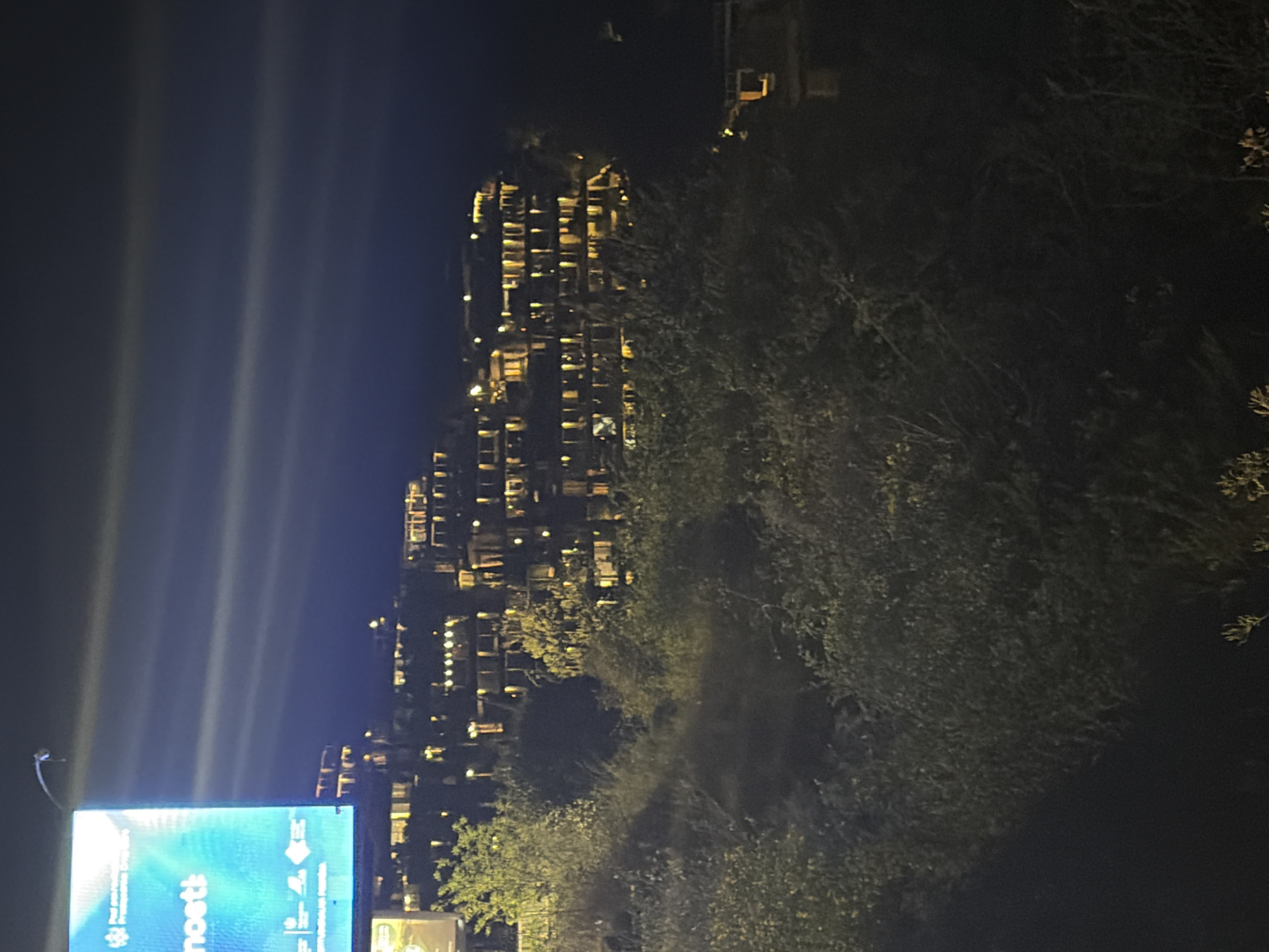 The image shows a night scene in Budva, Montenegro, featuring an illuminated advertising billboard in the lower left corner. In the background, a multi-story building with numerous lit windows stands against the night sky. Trees and greenery are visible on the right side of the frame, obscuring some of the view. The lighting and shadows suggest it is night. The image is rotated vertically.
