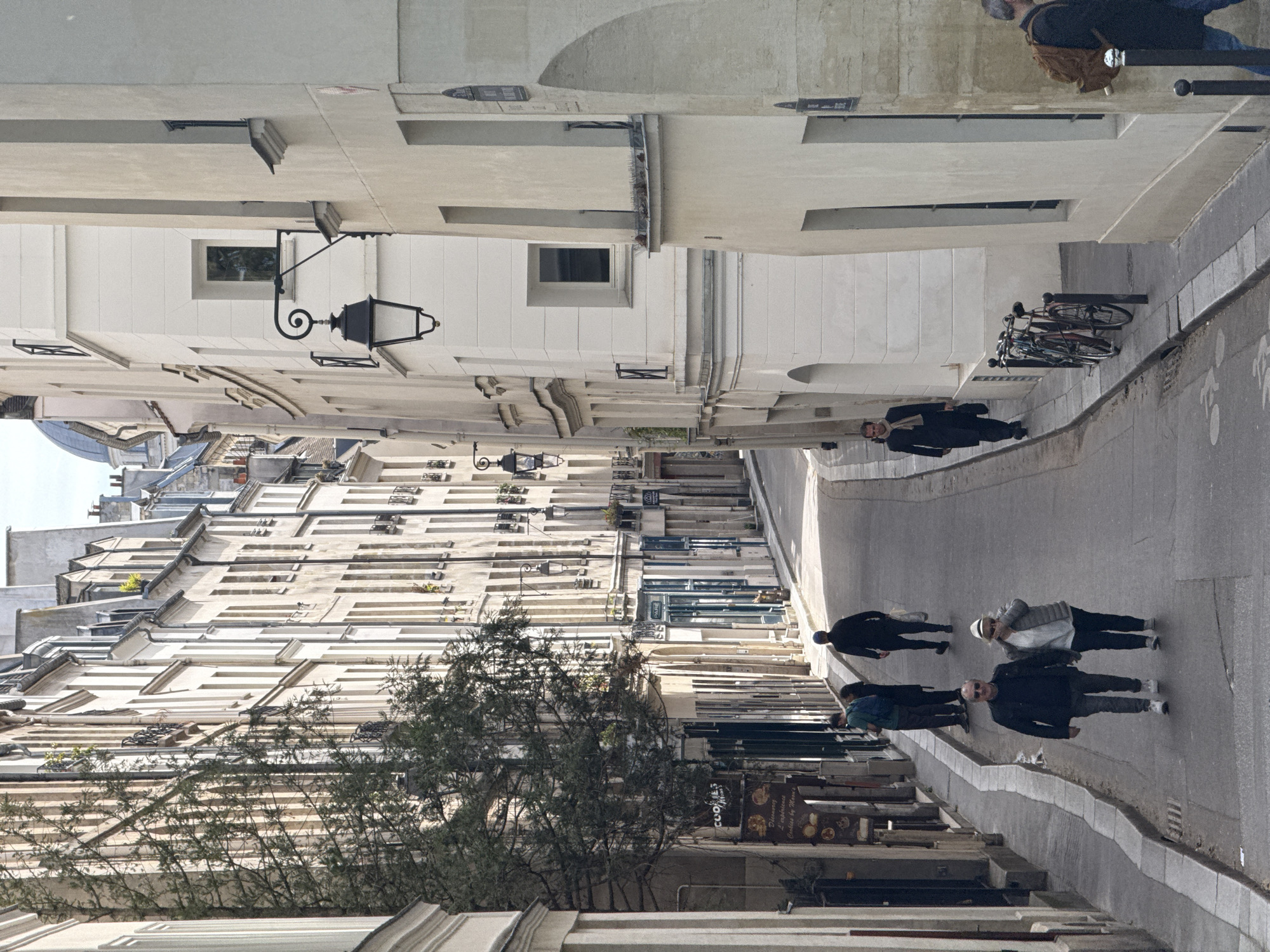 A narrow street in Paris, France, lined with buildings of various heights and architectural styles, stretches into the distance. Several people are walking on the right side of the street, and bicycles are parked along the curb. A street lamp with a curved arm is attached to a building on the left. A tree partially obscures the lower part of the image. Signs and awnings are visible above shopfronts. The sky is overcast.