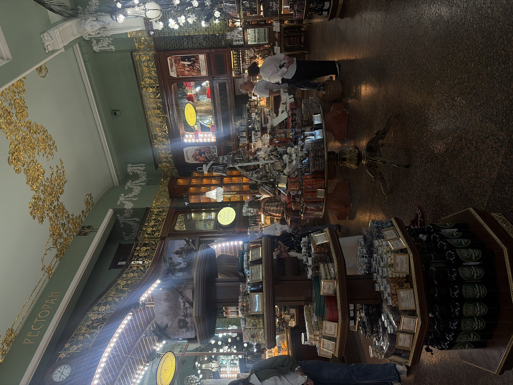The image depicts the interior of a well-decorated confectionary shop, likely in Saint Petersburg, Russia, suggested by the Cyrillic text "РЕСТОРАН" on the ceiling and "КОНДИТЕРСКИЕ ИЗДЕЛИЯ" on a yellow sign. The store features ornate gold detailing on the ceiling and railings, green walls, and chandeliers. Several display shelves are filled with various confectionery items in jars and boxes, as well as bottled goods on the lower level. People are seen browsing and interacting within the shop. One woman in jeans and a green jacket is on the left, and two people, a man and a woman, are in conversation in the middle right of the frame. The flooring is a dark, textured material.