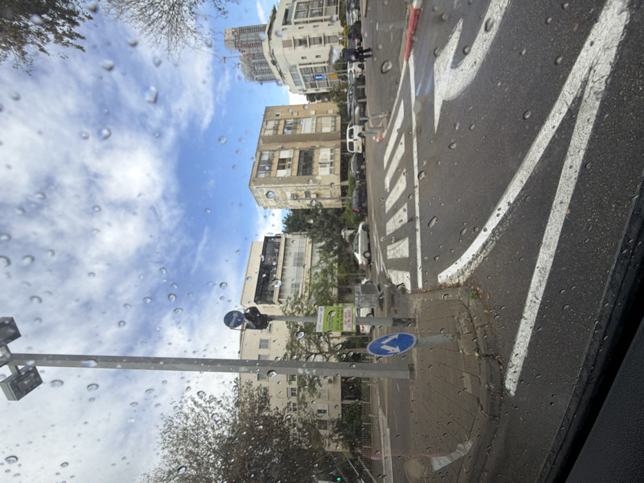 The image depicts an urban intersection in Tel-Aviv, Israel, specifically at the junction of Nordau Boulevard and Weizmann Street, observed through a rain-streaked windshield. The sky is partly cloudy with significant patches of blue. The asphalt road surface is wet, showing white painted road markings, including a crosswalk and a right-turn arrow. A red curb marking is visible on the right.
In the left foreground, a tall, grey utility pole supports multiple lights and a black traffic signal head. Below it, on a shorter pole, is a blue circular road sign with white arrows indicating "pass either side." Further back, green and white posters with Hebrew text are affixed to a pole. A silver sedan is partially visible to the left. The sidewalk on the left is paved.
To the left-center, a light-colored multi-story building with balconies is visible. In the center-right, an older, multi-story building with a weathered, discolored facade stands. To its right, a newer, light-colored building with multiple balconies and modern design elements is present. In the far background, a tall building under construction, topped with a crane, rises against the sky.
Numerous parked cars of various models and colors line the street on the right side. A single individual, dressed in dark clothing, is visible walking on the far right sidewalk, near the parked vehicles and the modern building. Green trees and foliage are scattered throughout the scene.