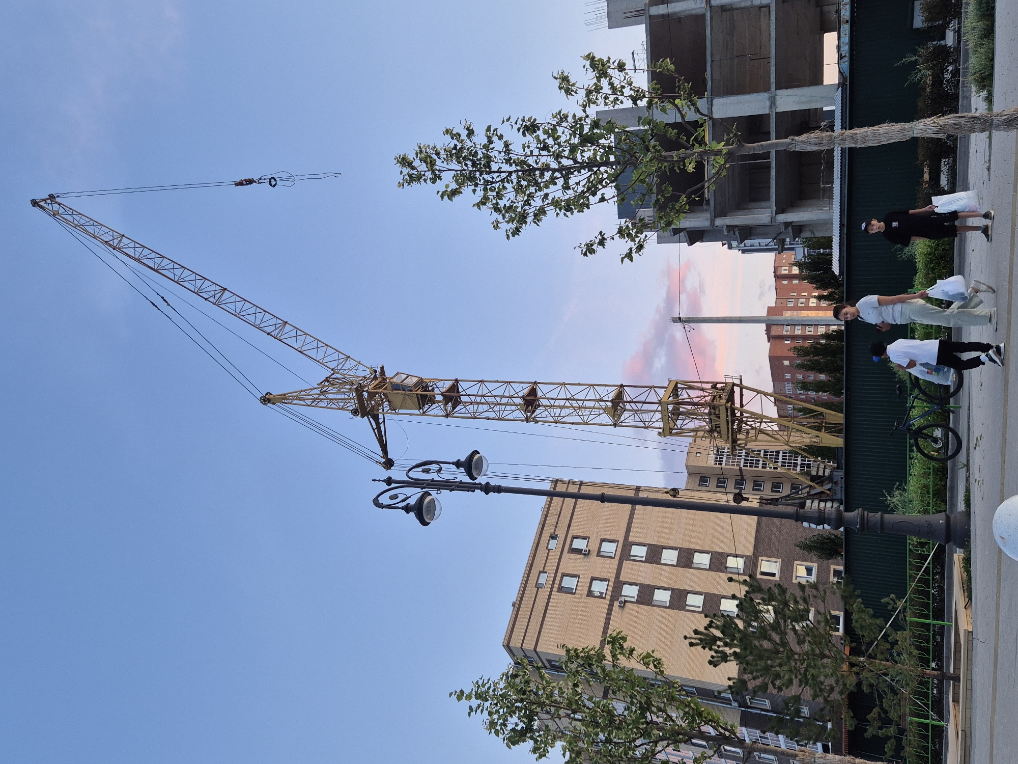 In Kokshetau, Kazakhstan, a construction site is visible under a clear sky with hints of pink and orange from the setting sun. A tall yellow tower crane dominates the scene, with its boom extending diagonally upwards. To the left, a light brown brick apartment building with numerous windows stands adjacent to a young tree. A vintage-style street lamp with two globes is positioned in the foreground. In front of a dark green fence, a bicycle is parked. Three individuals are walking on a paved walkway. A woman in a light-colored t-shirt and pants, carrying a white shopping bag, walks with a young boy in a white t-shirt and black pants, who also has a shopping bag. Behind them, a boy in a black t-shirt and shorts, carrying a white shopping bag, walks. Further back, an unfinished concrete building with visible rebar is under construction.