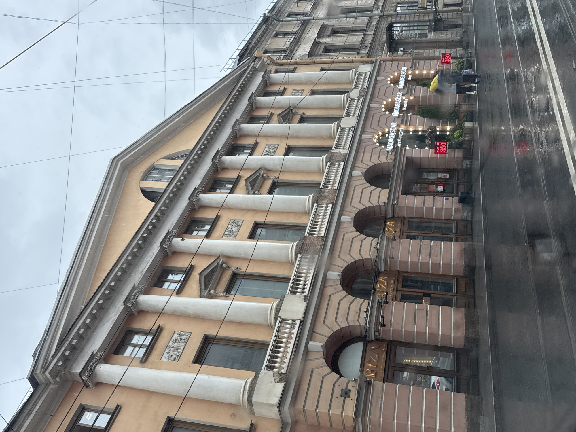 The image shows a rain-slicked street in Saint Petersburg, Russia, with a grand, peach-colored building dominating the foreground. This building features classical architecture with large white columns, arched doorways on the ground floor, and a balustrade along the second story. Several businesses are visible on the ground floor, including "BIZA" and "MADAGASCAR restaurant karaoke bar". Three red signs with the text "все по 350" (everything for 350) are displayed outside the Madagascar establishment. Two individuals, one holding a yellow umbrella and the other a black umbrella, are walking on the wet sidewalk in front of the Madagascar establishment. The street reflects the overcast sky and the surrounding buildings, indicating wet conditions. The sky is overcast and grey.