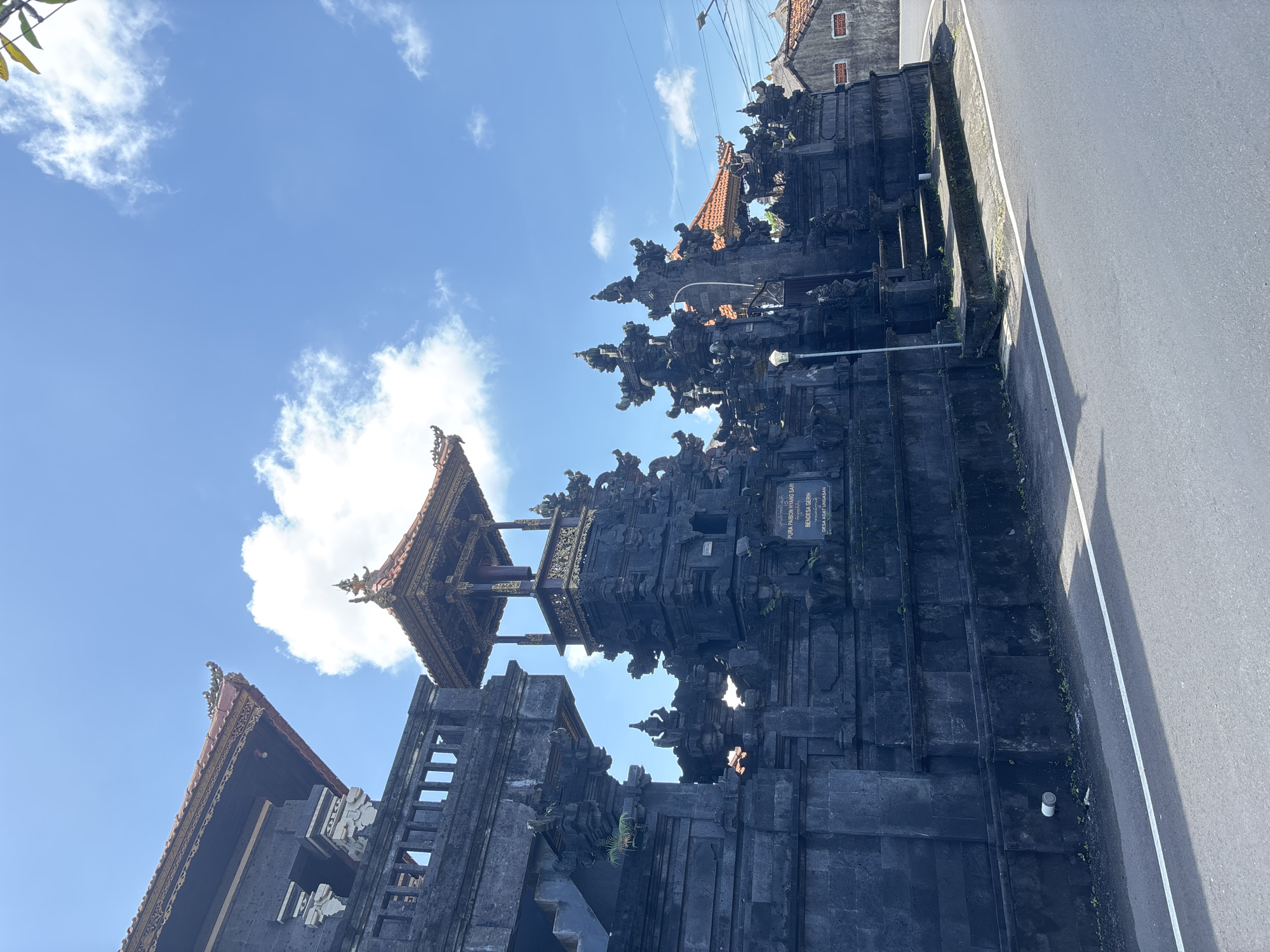 The image shows a traditional Balinese gate structure alongside a road in Kuta Selatan, Indonesia. The gate is constructed from dark stone, featuring intricate carvings and tiered roofing elements. A smaller building with a tiled roof is visible to the left of the gate. A road with a white painted line runs adjacent to the gate. The sky is blue with scattered white clouds.