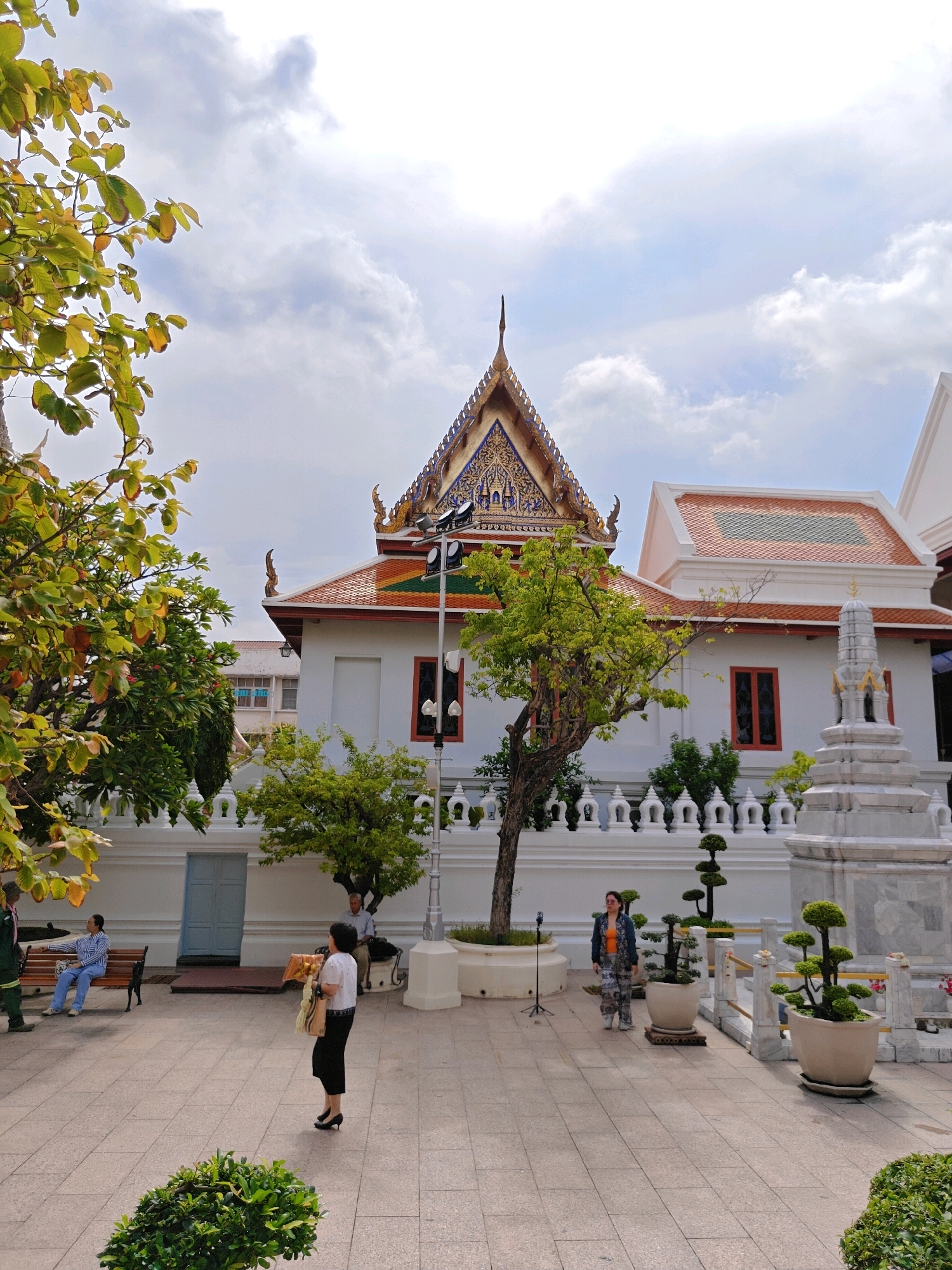 This image shows a view of Wat Arun, a temple in Bangkok, Thailand. In the foreground, there is a woman in a white shirt and black skirt holding a flower garland and a bag. To her left, a man in a blue and white striped shirt and blue pants sits on a bench. Behind the man, there is a white wall with decorative elements and a blue door.
In the middle ground, a traditional Thai temple building with a tiered roof is visible. The roof is adorned with intricate carvings and golden embellishments. Trees with green foliage are planted around the temple.
In the background, a clear sky with scattered clouds can be seen. The overall scene depicts a serene temple courtyard, with people engaging in various activities within the temple grounds. The architectural style and the presence of Buddhist iconography are characteristic of Thai temples.