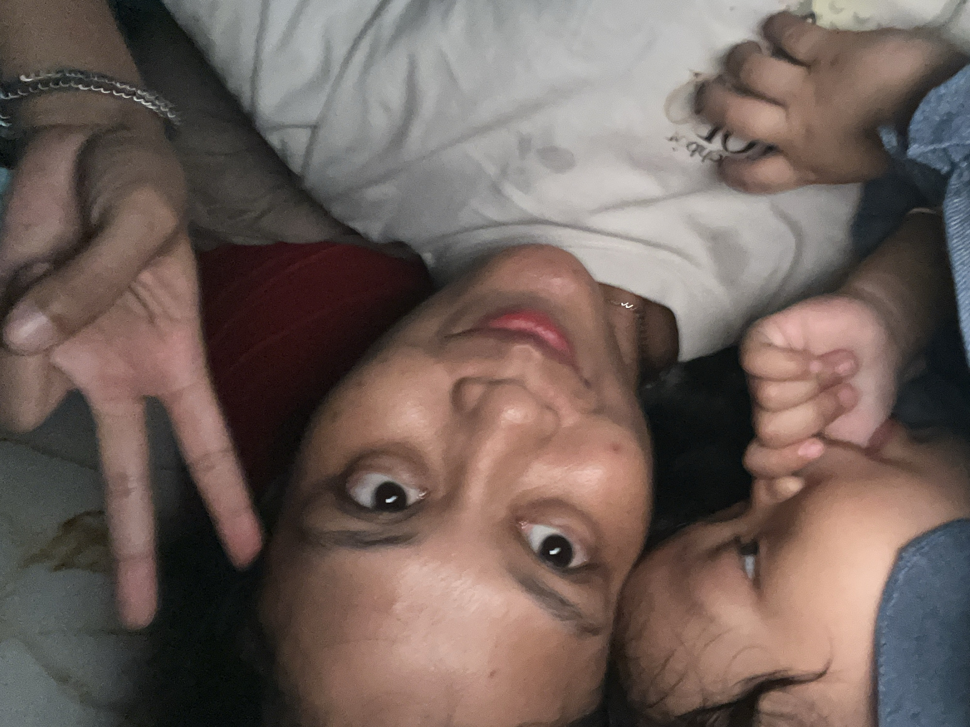 In a close-up, overhead shot, a young adult with dark hair and brown eyes is lying down, looking upwards at the camera. Their left hand is raised in a peace sign, with fingers spread. They are wearing a white t-shirt. Next to them, a baby is lying down, with their face partially obscured. The baby's hand is in their mouth. The background is blurry, but appears to be a bed with white and red bedding. The image appears to have been taken indoors in a residential setting in Kecamatan Pati, Indonesia.