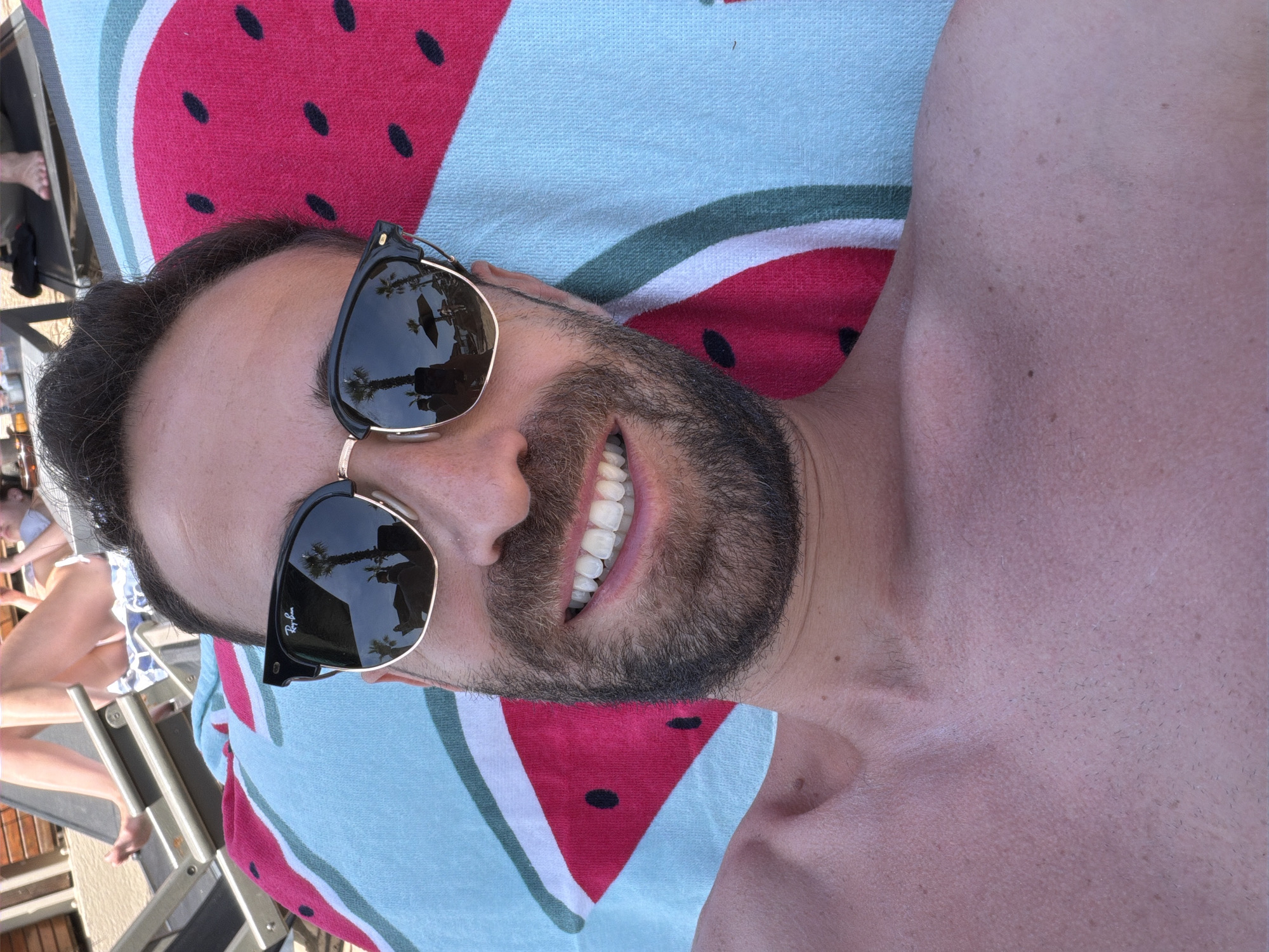 A man with a beard and dark hair is captured in a close-up, smiling broadly at the camera while lying down outdoors. He is wearing black and gold-framed Ray-Ban sunglasses, which reflect palm trees and a bright sky, suggesting a sunny, resort-like environment. He is shirtless, revealing his chest and collarbone, and is resting on a vibrant beach towel with a watermelon pattern in red, light blue, and white. In the blurry background to the left, parts of another person, likely a woman in a white bikini, and a lounge chair are visible. The scene conveys a relaxed, leisurely atmosphere, characteristic of a vacation or holiday in Alexandroupolis, Greece, during daytime.