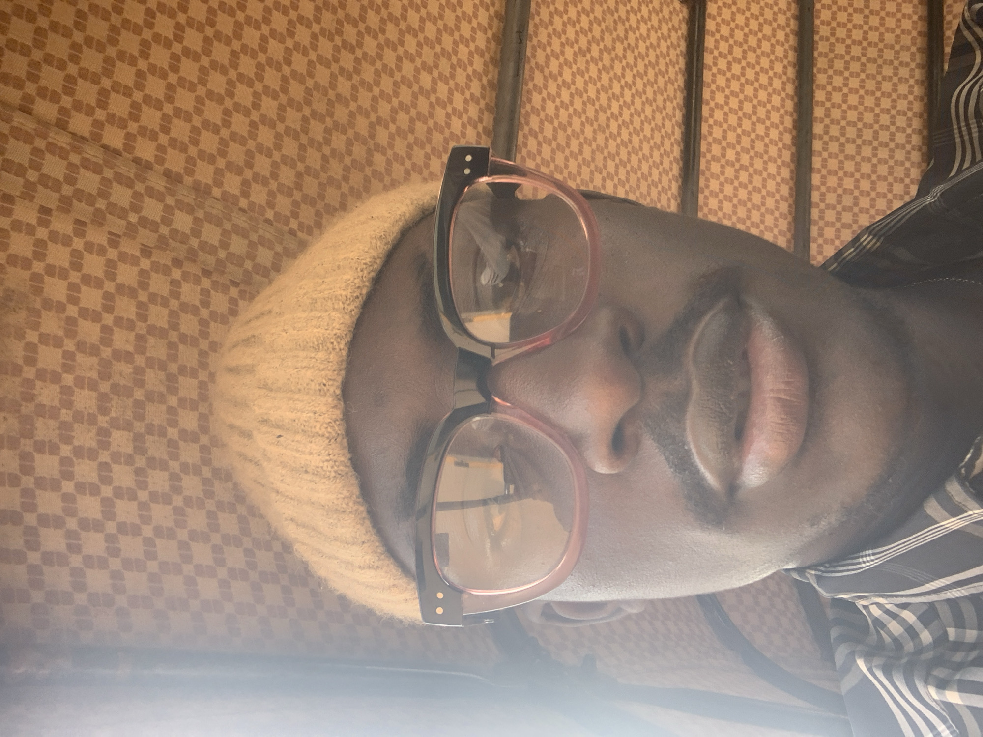The media file is a close-up, rotated image of a person's face. The person is wearing a light-colored beanie and large, rectangular eyeglasses with pinkish-brown frames. Their lips are slightly parted, and they appear to be looking upwards. The background is a textured fabric with a repeating diamond pattern in shades of brown and beige. The lighting suggests it might be daytime, with a strong light source coming from the lower right of the frame, creating a bright glare that obscures some details. The person is wearing a dark, checkered shirt, partially visible on the right side of the image. The setting appears to be indoors, possibly on a piece of furniture covered with the patterned fabric. There are no visible text elements in the image. The image does not provide explicit cues about the specific activity or event, but the pose and close-up framing suggest a casual, possibly selfie-style photograph.