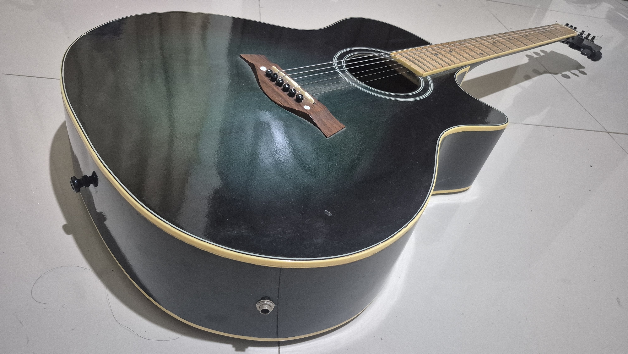 An acoustic guitar with a black and green gradient finish lies on a tiled floor. The guitar is positioned at an angle, with its headstock pointing towards the right. The body of the guitar features a wooden bridge with six black pins holding the strings. The soundhole is surrounded by a white rosette. The neck of the guitar is made of light-colored wood, and its fretboard is visible. The headstock has tuning pegs. The guitar appears to be an electric-acoustic model, as there is an output jack on the bottom of the body. There is also a strap button on the side of the guitar. The tiled floor has a light gray color and a subtle pattern. The lighting in the scene is artificial, casting shadows from the guitar onto the floor. The image was taken in Bojonegoro, Indonesia.
