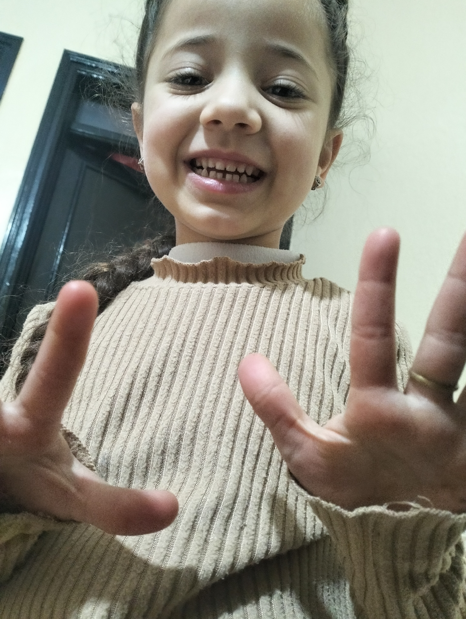 A young girl with brown curly hair pulled back in a loose braid smiles broadly at the camera, holding up both hands. The skin on her face and hands is fair. She wears small earrings. The girl is wearing a beige, ribbed sweater with a ruffled neck and wrist cuffs. The background includes a beige wall and a partially visible dark-colored doorframe. The image's location and specific setting within Ouaouizert, Morocco, are not discernible from the provided visual information.