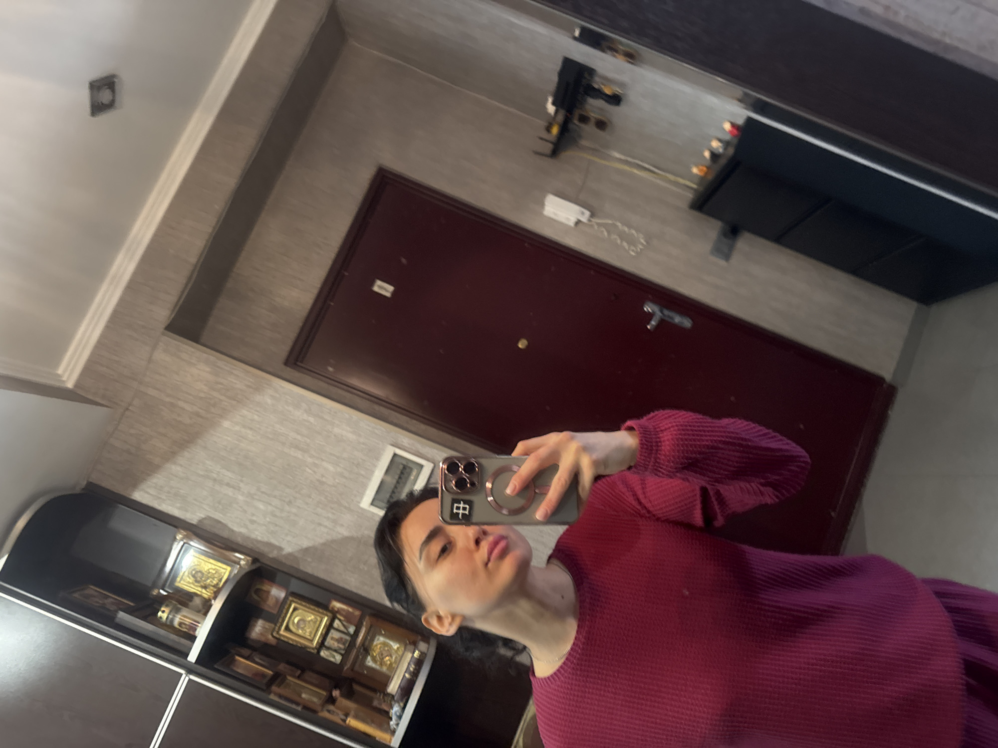 A young adult female is depicted in a mirror selfie taken within a residential hallway in Tbilisi, Georgia. She has dark hair and is wearing a magenta-colored, long-sleeved waffle-knit top. Her left hand holds a dark-colored smartphone, visible with a prominent camera array and a circular magnetic accessory on its back. The phone is positioned to capture her reflection.
The background reveals interior features typical of a Georgian home. To the left of the subject, a dark brown display cabinet with glass shelves is filled with multiple framed Orthodox Christian icons, predominantly featuring golden and brown hues. A glass votive candle holder is also visible on one shelf. The wall directly behind the subject and extending to the right features light beige/grey textured wallpaper.
To the right of the subject, a dark red entrance door is visible, equipped with a silver handle, a circular brass peephole, and two small white rectangular labels affixed near the top. A white wired intercom unit is mounted on the wall to the left of the door. Below the intercom, a tall, narrow, black shoe cabinet with multiple compartments stands, topped with a few small decorative items. Above the shoe cabinet and to the right of the intercom, a black wall-mounted shelf or key holder is affixed, from which keys and possibly sunglasses are hanging. A white rectangular air vent is positioned high on the wall above the door. The ceiling is white with white crown molding, featuring a square recessed light fixture.