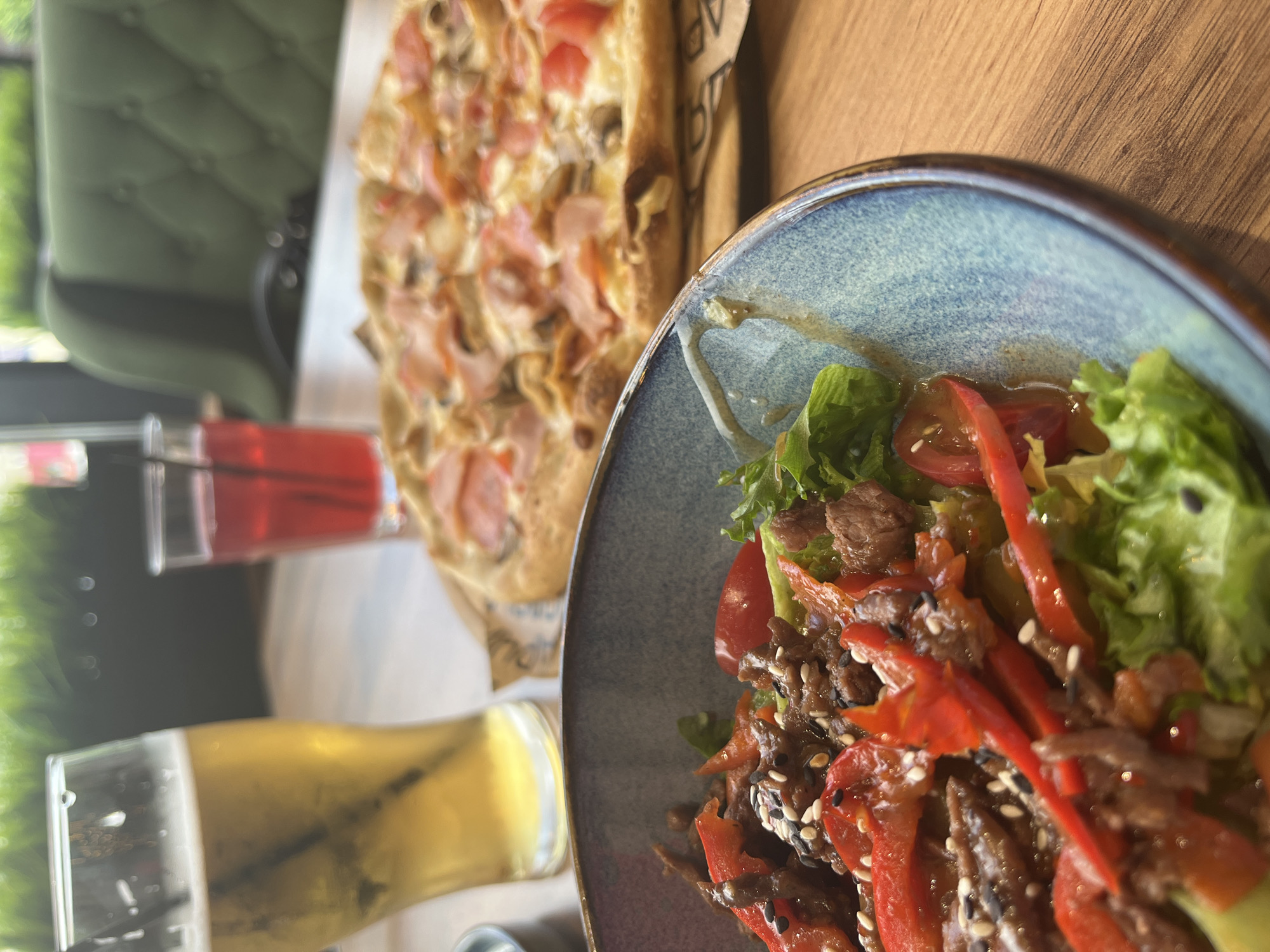 In Izhevsk, Russia, a close-up shot captures a dining table at a restaurant, featuring a meal set for one. In the foreground, a blue ceramic bowl overflows with a vibrant salad. The salad consists of a base of crisp green lettuce, topped with thinly sliced red bell peppers, pieces of cooked meat, and cherry tomato halves. The dish is sprinkled with white and black sesame seeds, and a light dressing glistens on the ingredients.
To the right of the salad, a partially visible pizza rests on a rustic wooden board. The pizza appears to have a thin crust, topped with melted cheese, slices of ham, and mushrooms, and is cut into several triangular slices.
Behind the food, two glasses stand on the table. The left glass is filled with a pale yellow beverage, likely beer, with a straw inserted. Next to it, a shorter glass contains a dark red liquid, possibly a fruit drink or juice, also with a straw.
The background is softly blurred, showing hints of the restaurant's ambiance. A plush, dark green upholstered chair is visible, along with out-of-focus greenery, suggesting an indoor setting with an outdoor view or decor. The table surface is a warm-toned wood grain. The lighting appears to be natural, with a slightly warm cast. The image is composed to focus on the food, with the background elements providing context without distracting from the primary subjects.