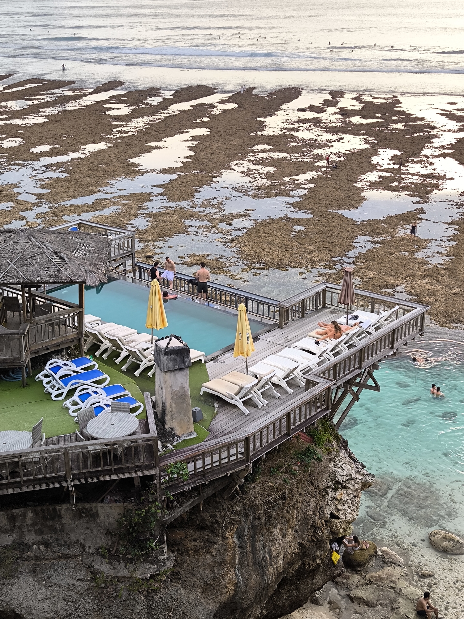This image captures a coastal scene likely in Uluwatu, Indonesia, displaying an elevated wooden structure jutting out over a rocky cliffside. On the structure's deck, there is an infinity pool where several people are present. Some individuals are at the edge of the pool, while others are swimming in the adjacent ocean. The deck is also furnished with lounge chairs and tables, and a thatched-roof gazebo is visible. The surrounding environment includes a shallow expanse of water-covered terrain and the ocean in the distance where people are surfing. The foreground features the rough surface of the cliff and a small beach area with people. The overall scene portrays a resort setting integrated with the natural coastal environment.