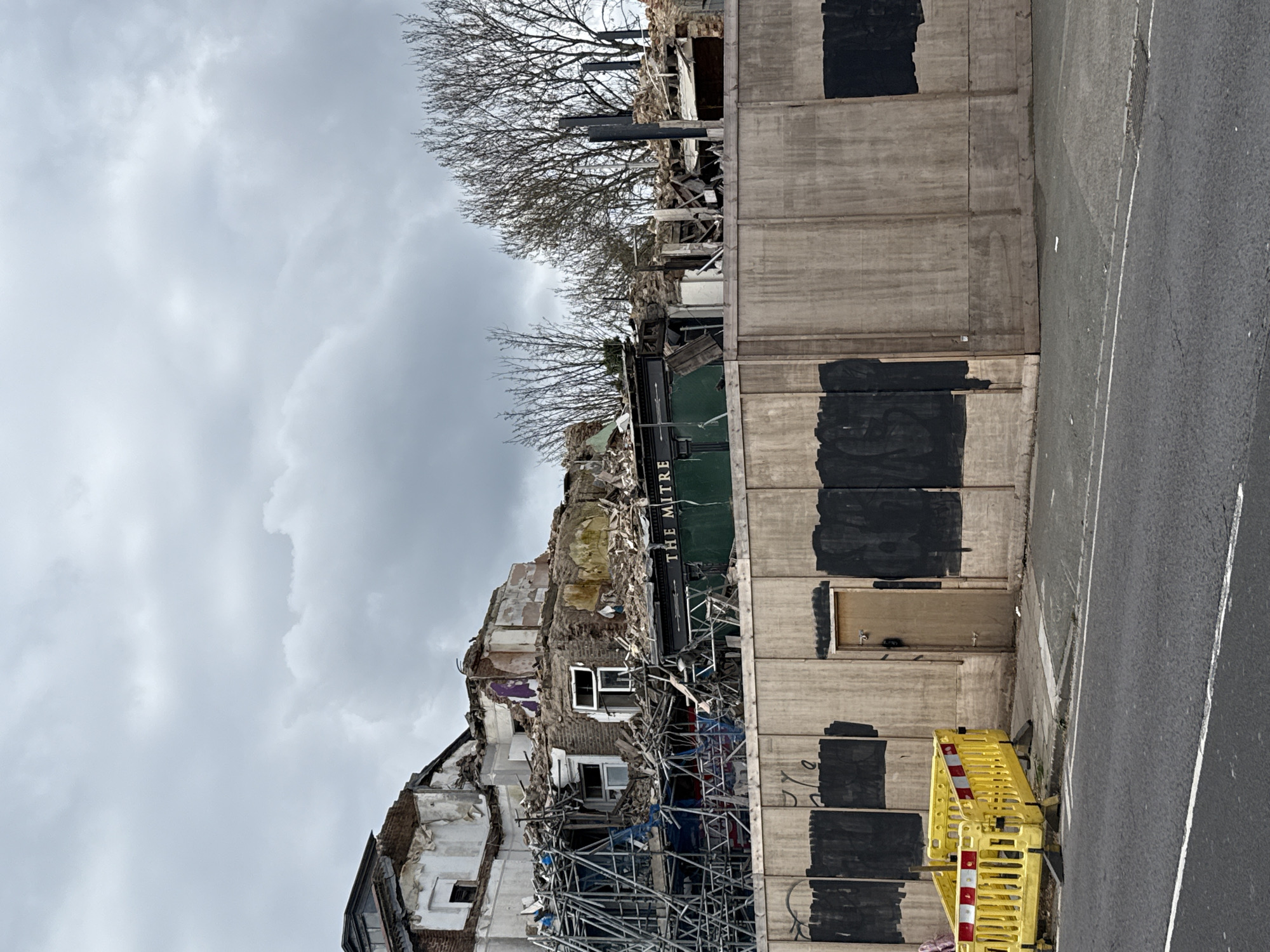 The image depicts the partially demolished remains of "The Mitre" pub in London, United Kingdom. The building's façade is severely damaged, with rubble, exposed brickwork, and scaffolding visible. A green sign reading "THE MITRE" hangs above the destruction. Adjacent to the pub is a concrete wall with several black rectangles painted over graffiti. At the base of the wall, there is a small wooden door and a yellow and red safety barrier. A gray asphalt road with white lane markings is present on the right side of the frame. The sky is overcast with gray clouds, and bare tree branches are visible above the demolished building.